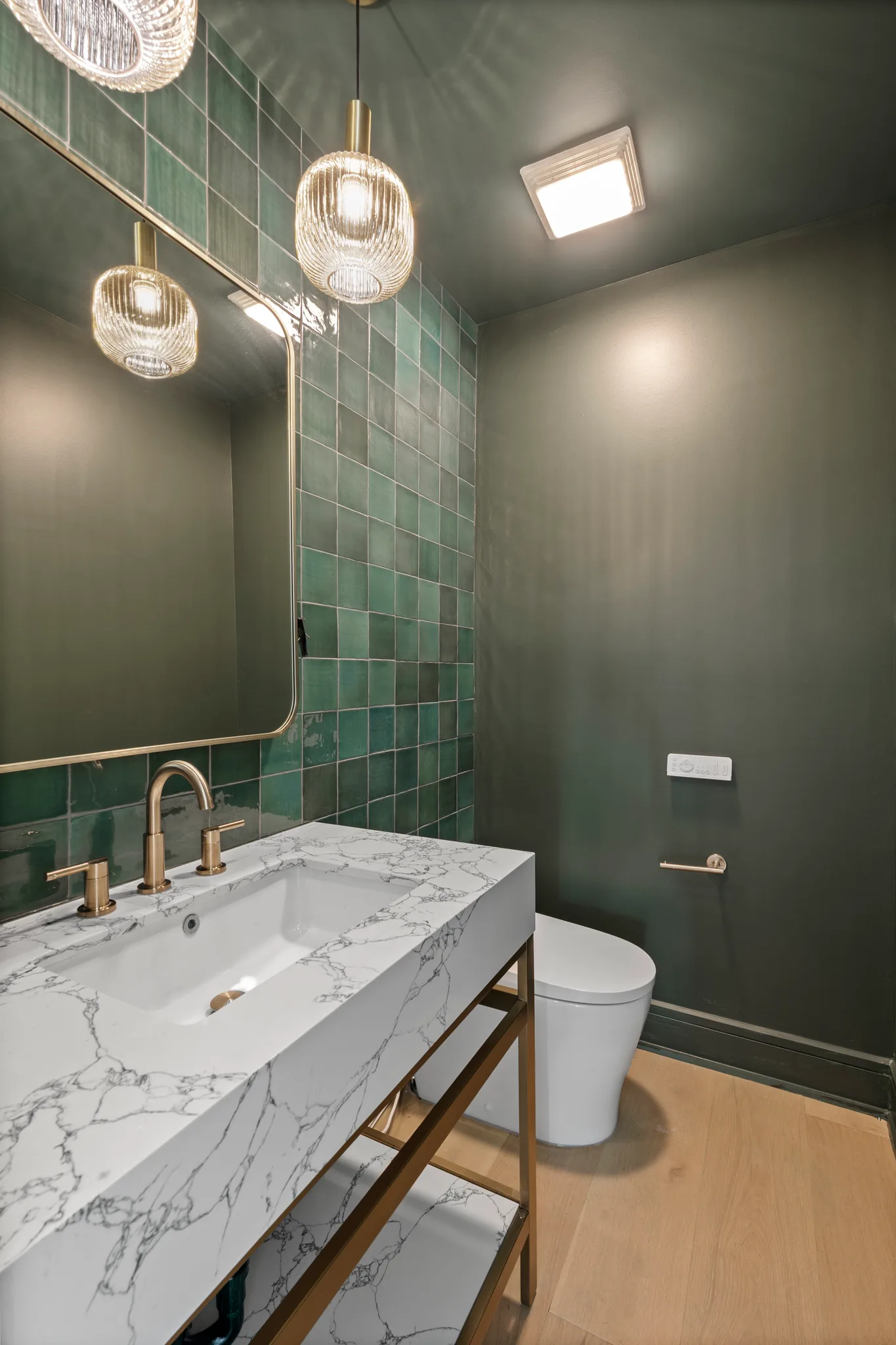 Powder room with a glossy green tile accent wall, marble-look trough sink on a brass stand, brass mirror and ribbed glass pendant lights