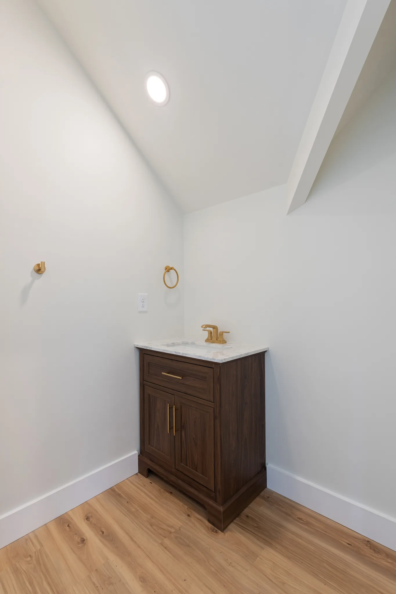 Small powder room with a dark walnut vanity, marble-look counter, brass faucet and towel ring under a sloped white ceiling