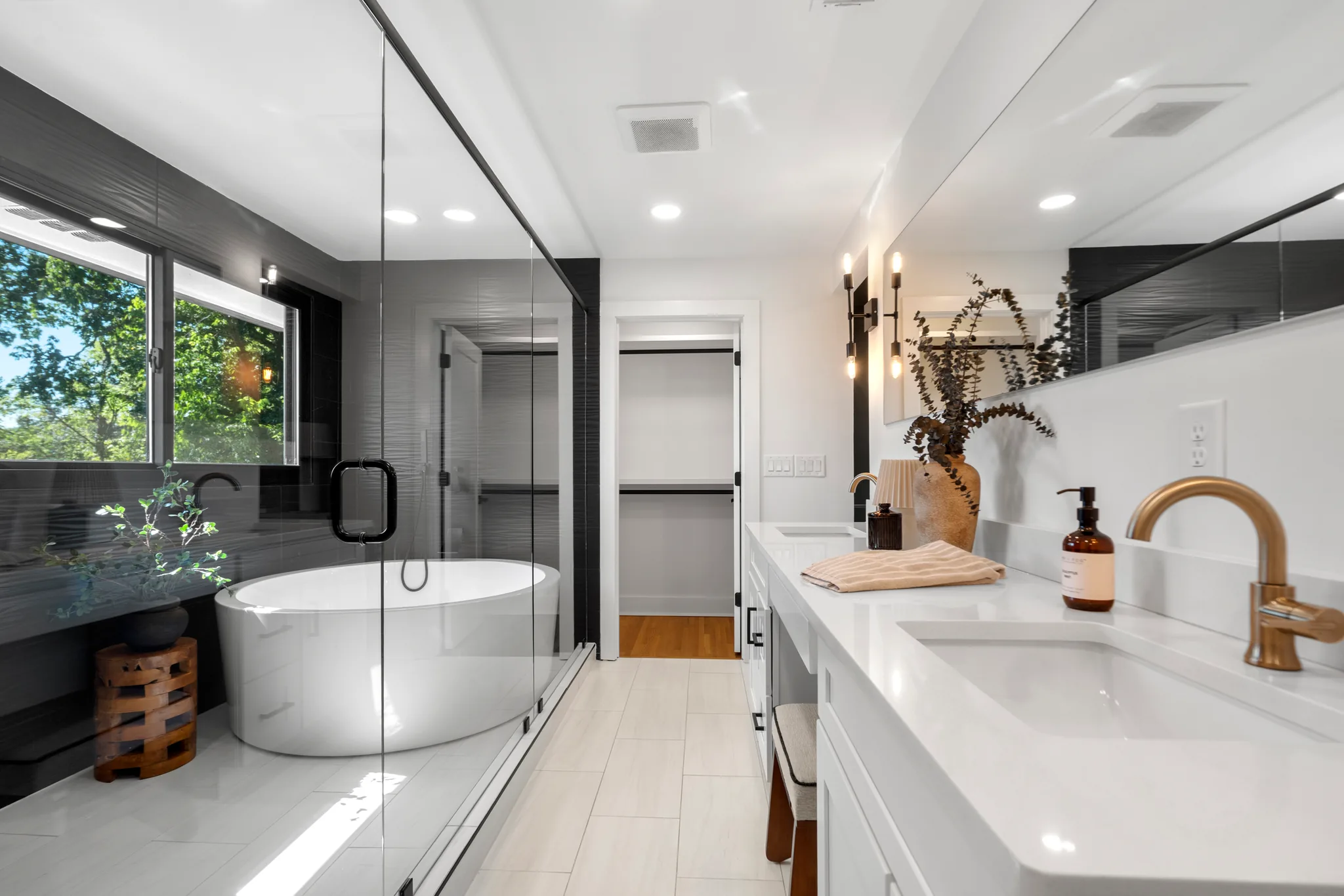 Primary bathroom with oval freestanding tub, glass shower enclosure, dark textured tile wall, white double vanity with brass faucets, and large-format floor tile