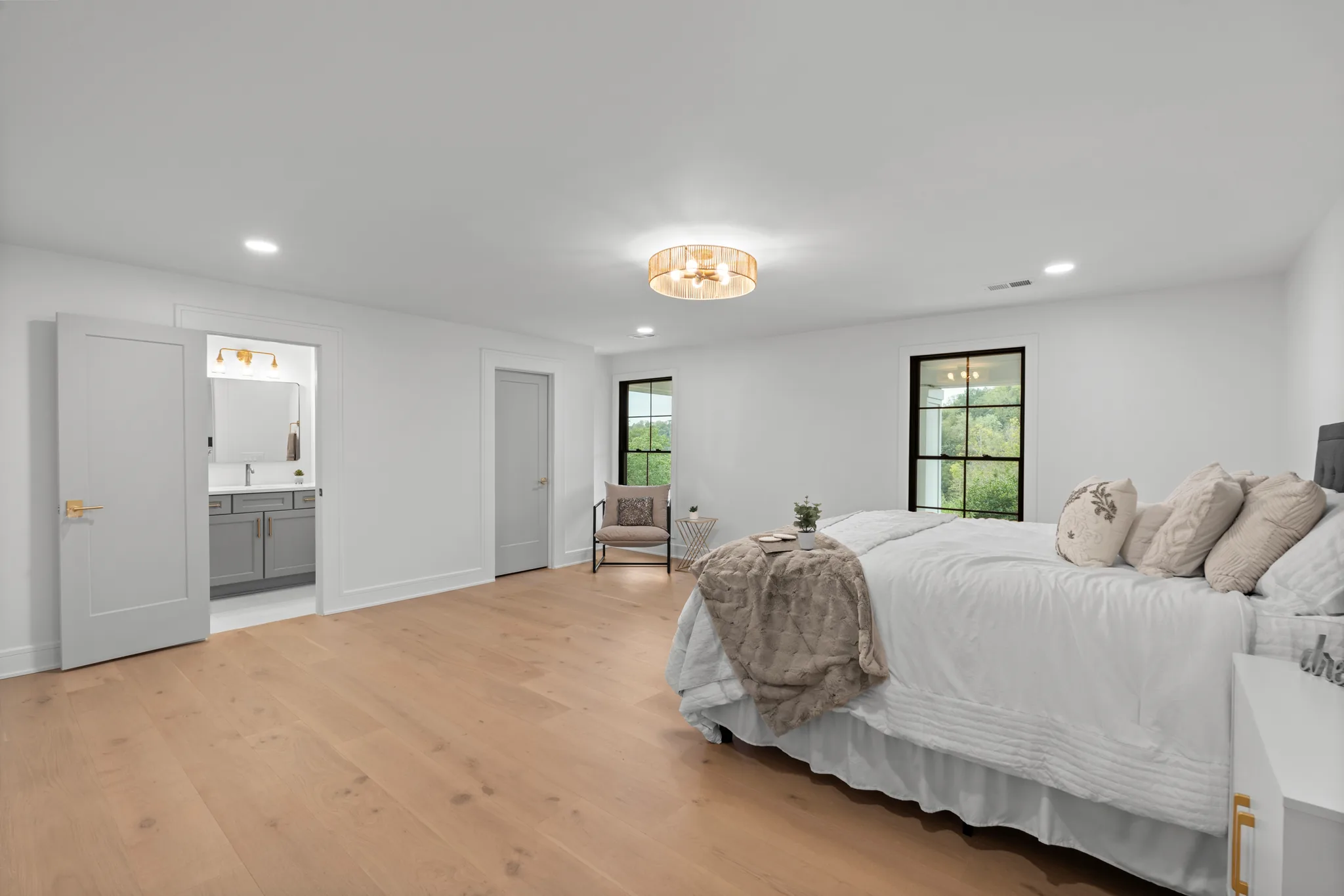 Primary bedroom with white walls, wide-plank oak floors, black-framed windows and an open doorway to a bath with a gray vanity