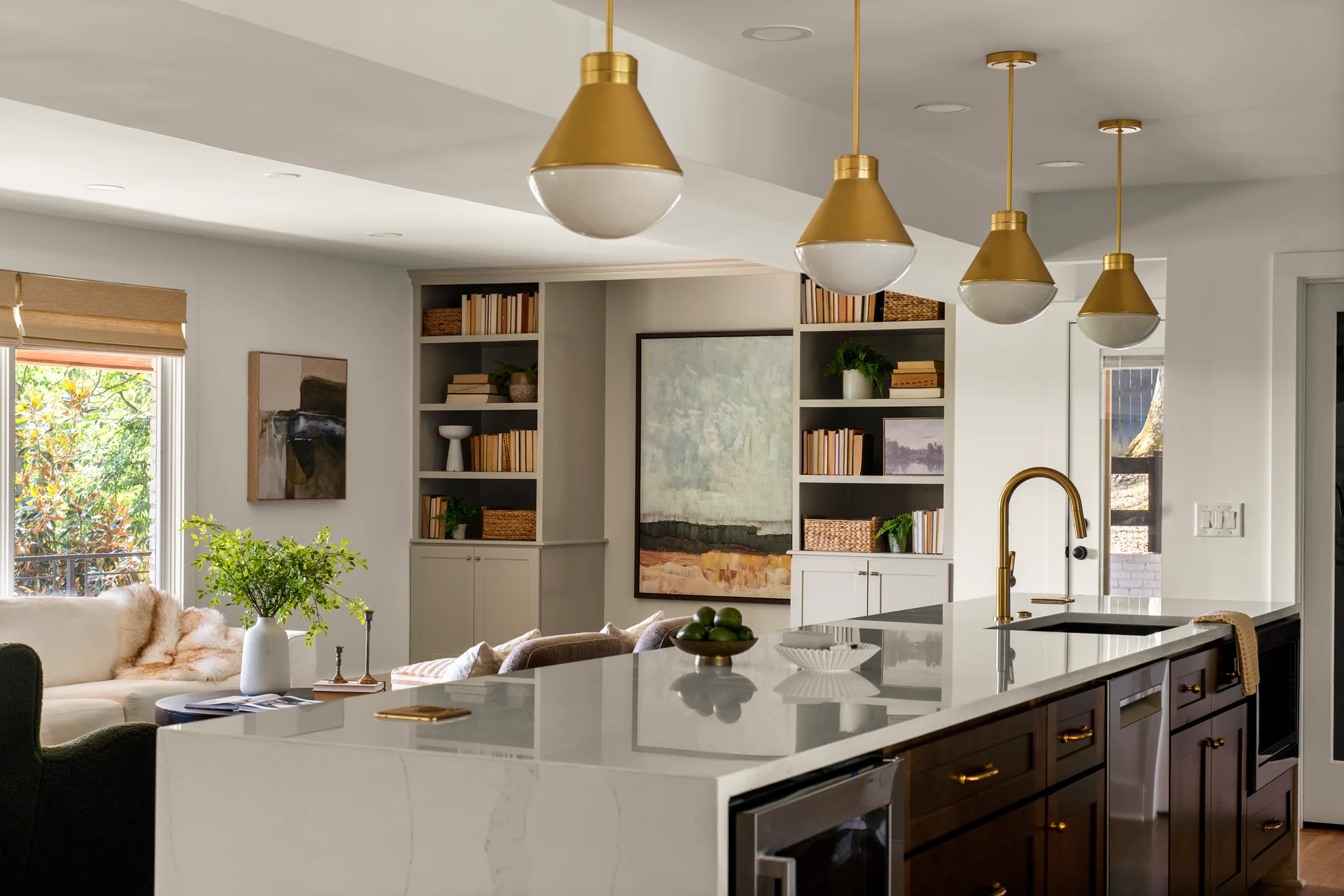 Kitchen island with white veined quartz waterfall countertop, dark wood cabinets, brass pendant lights overhead and an adjoining living area with built-in shelves