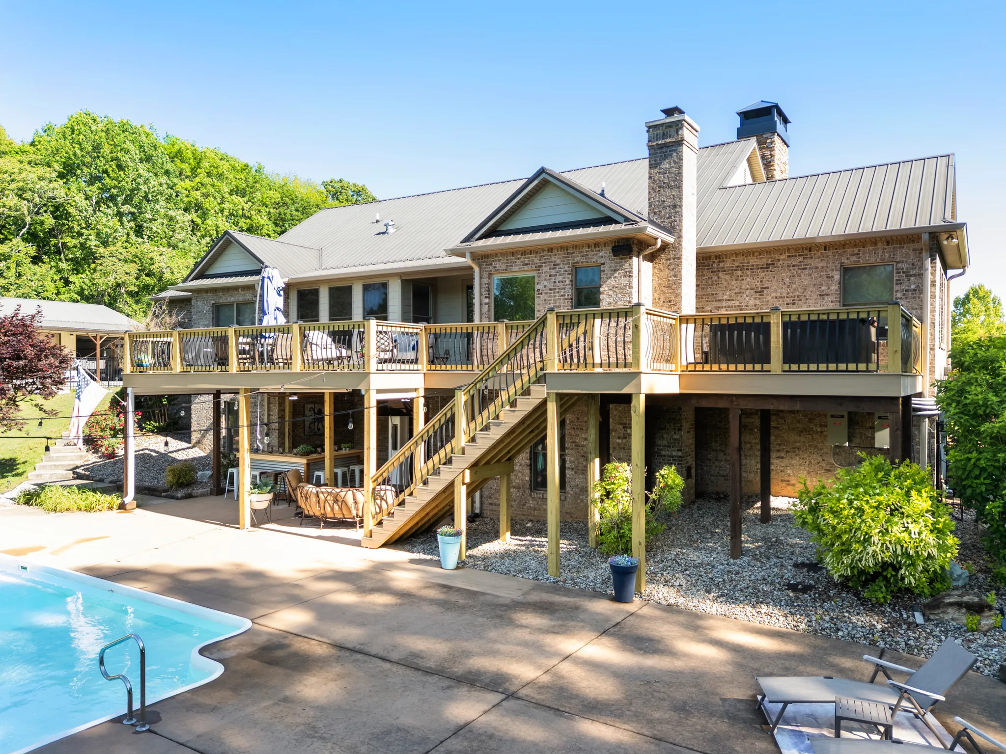 Raised wood deck with black metal balusters spanning the rear of a brick home with metal roof, stairs down to a concrete pool patio