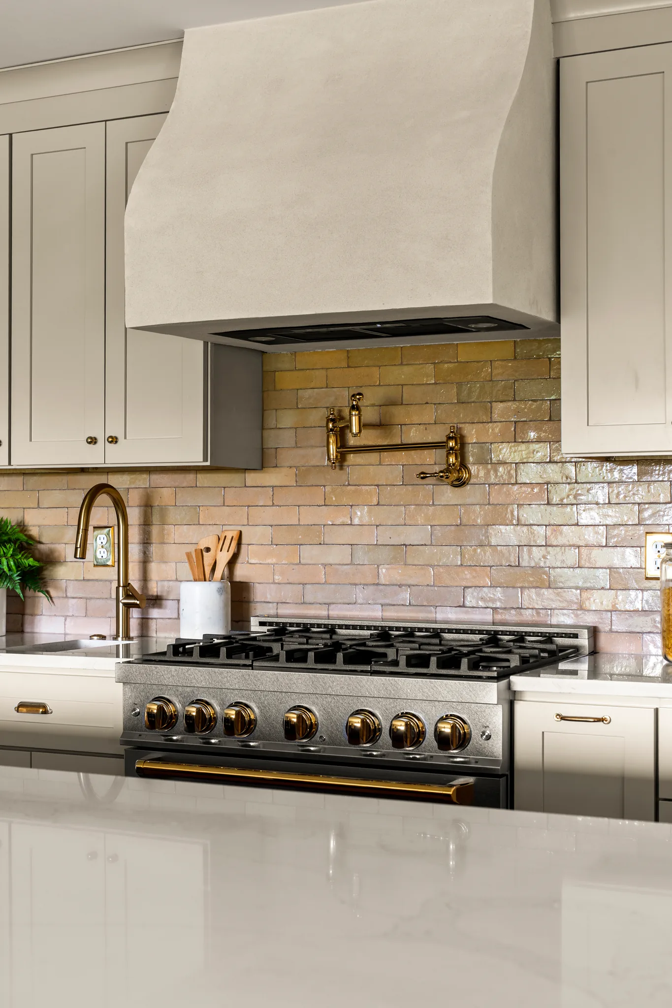 Stainless range with brass knobs set under a curved plaster hood, with an iridescent glazed brick tile backsplash and cream shaker cabinets