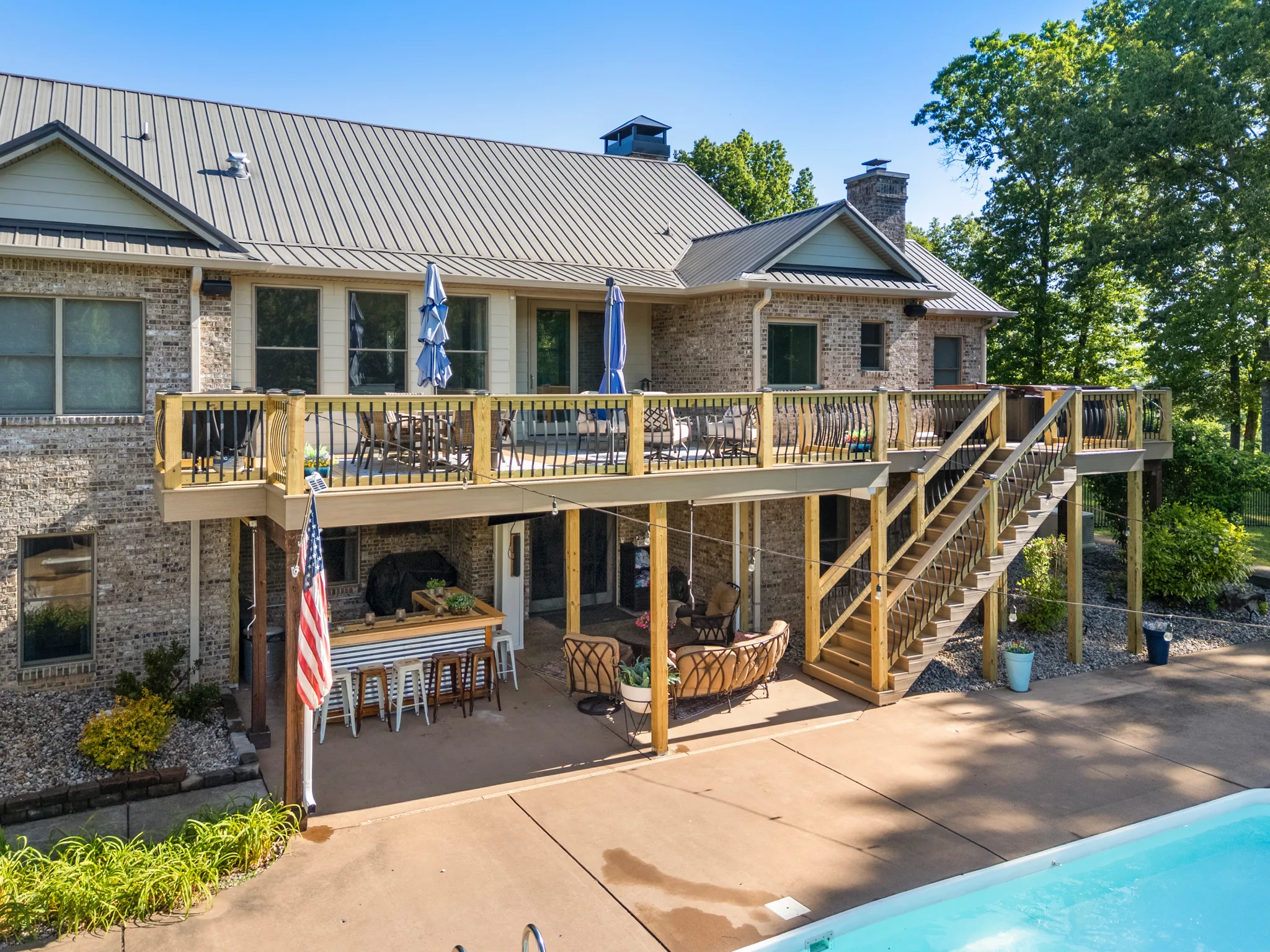 Raised wood deck across the back of a brick home with metal roof, open stairs to a concrete pool patio below