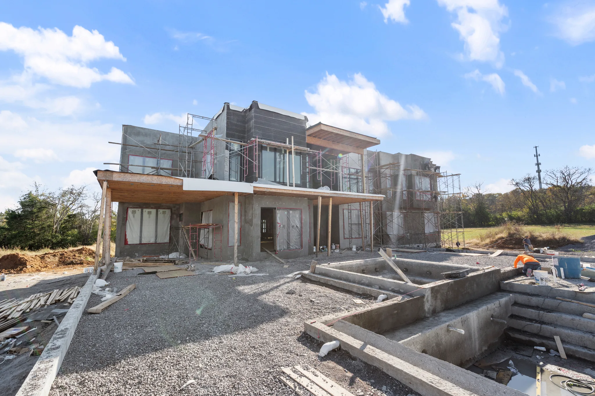 Two-story modern home under construction with scaffolding, wood soffits and unfinished walls above concrete forms set into a gravel yard