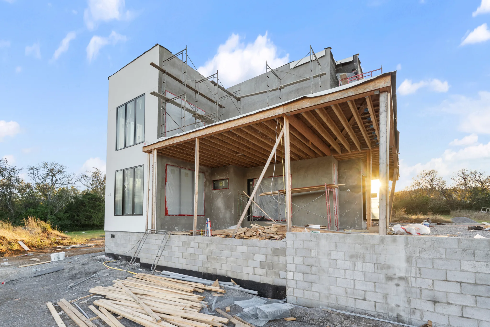 Modern two-story home under construction with exposed porch rafters, stucco walls, scaffolding and a concrete block retaining wall in front