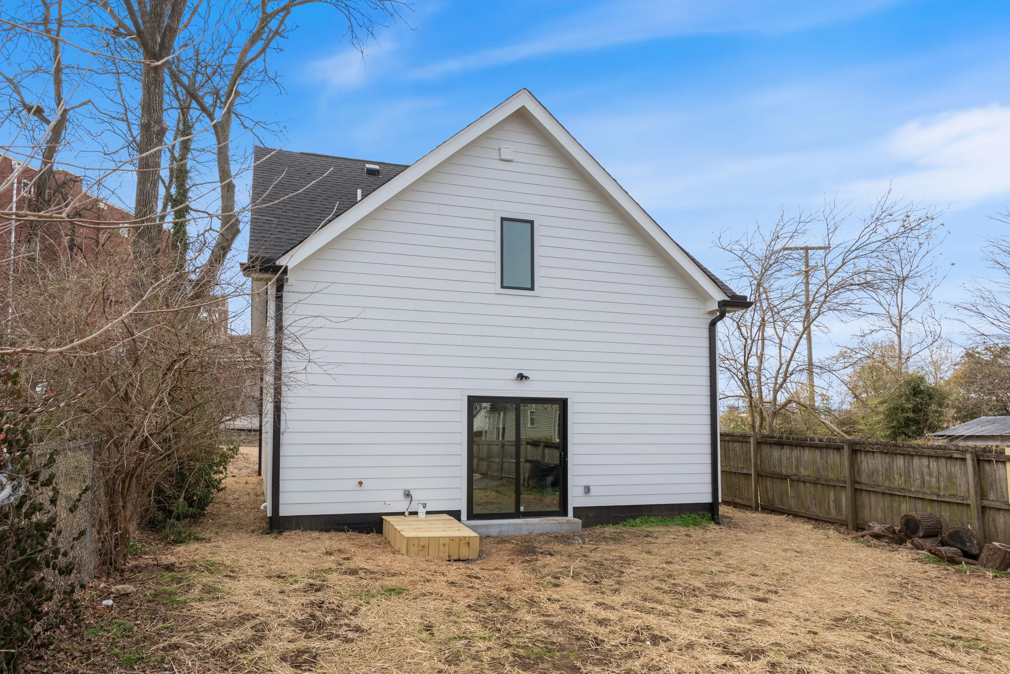 Rear elevation of a white lap-sided home with a steep gable, black-framed sliding glass door, tall narrow window and wood step platform