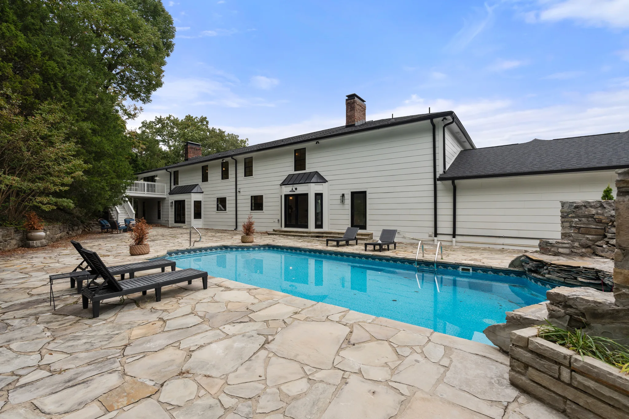 Rectangular swimming pool set in a wide irregular flagstone patio behind a long white home with black windows and two brick chimneys