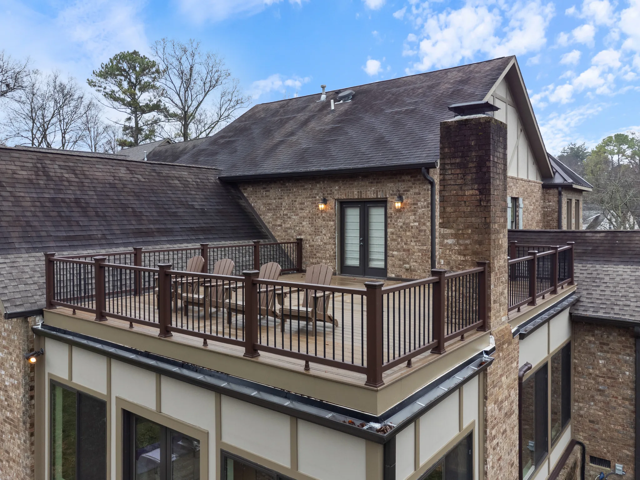 Elevated deck with dark wood railing and black balusters set between brick gables, with Adirondack chairs facing French doors