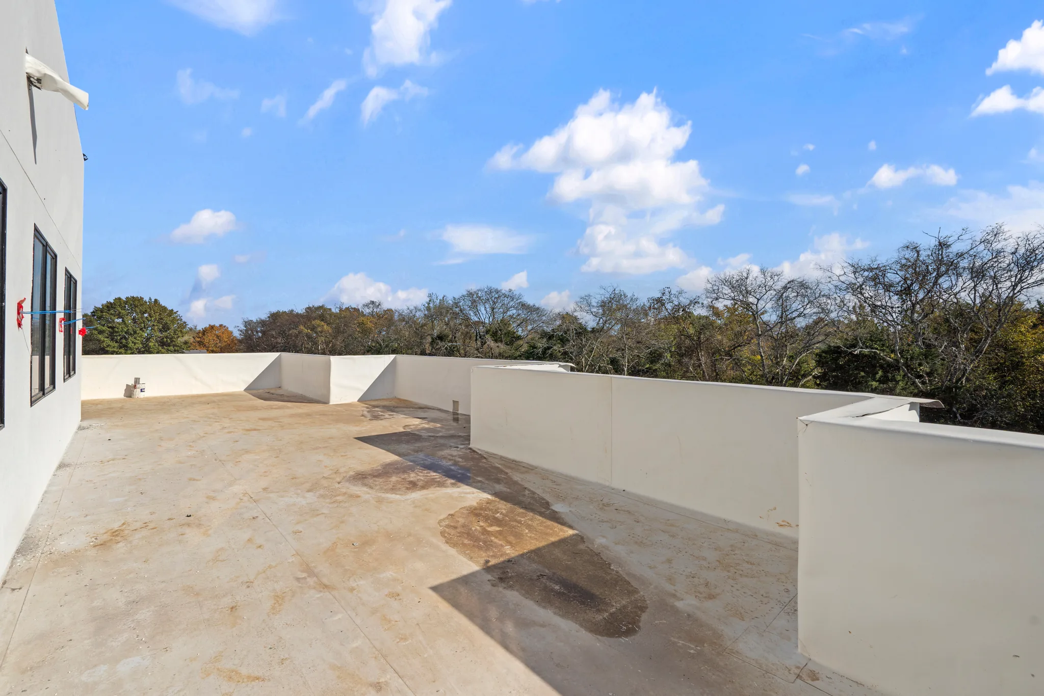 Flat rooftop deck under construction with white stucco parapet walls, bare concrete surface, dark window wall at left and treetops beyond