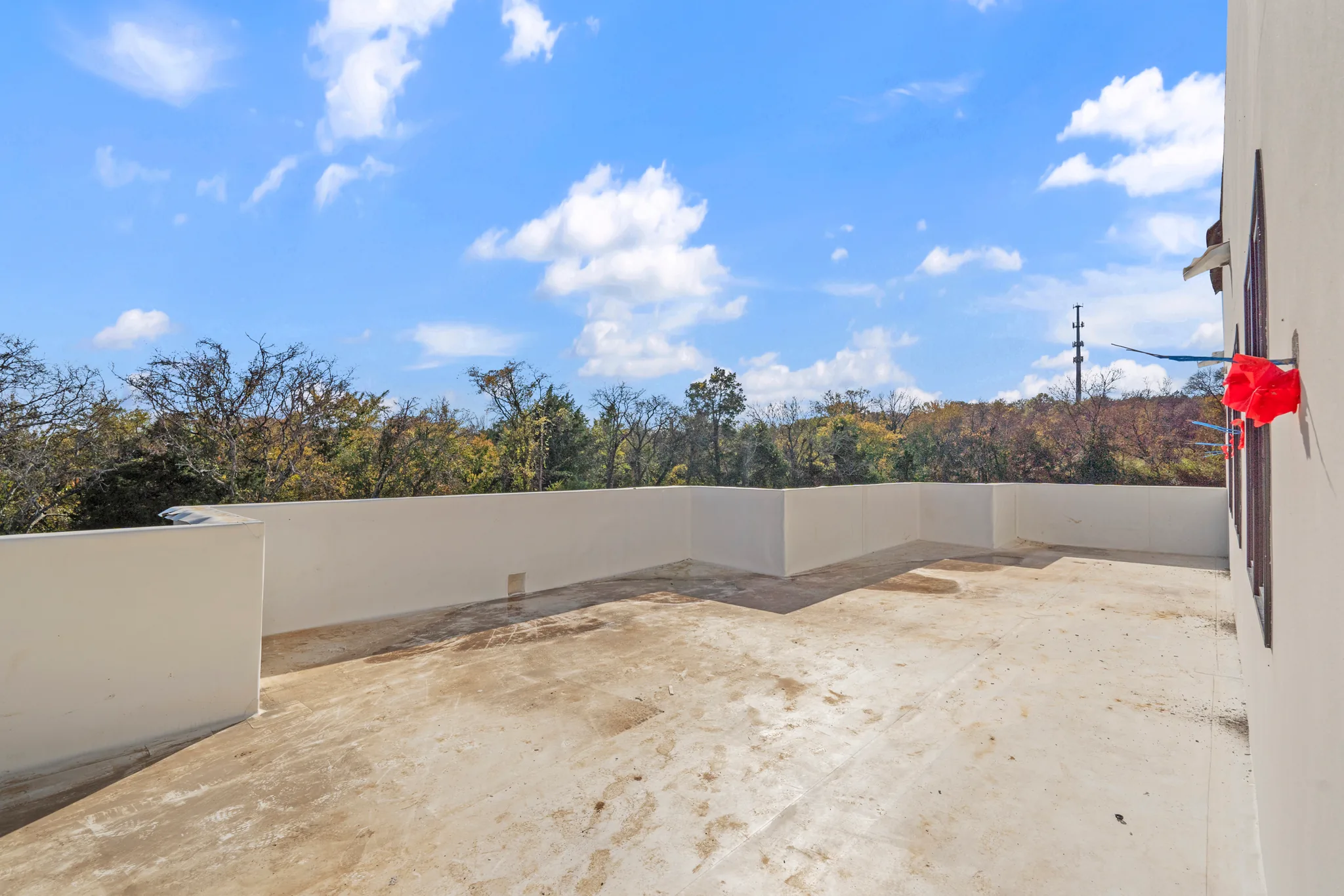 Rooftop terrace under construction with a white stucco parapet wall, bare concrete floor and treetops under a blue sky