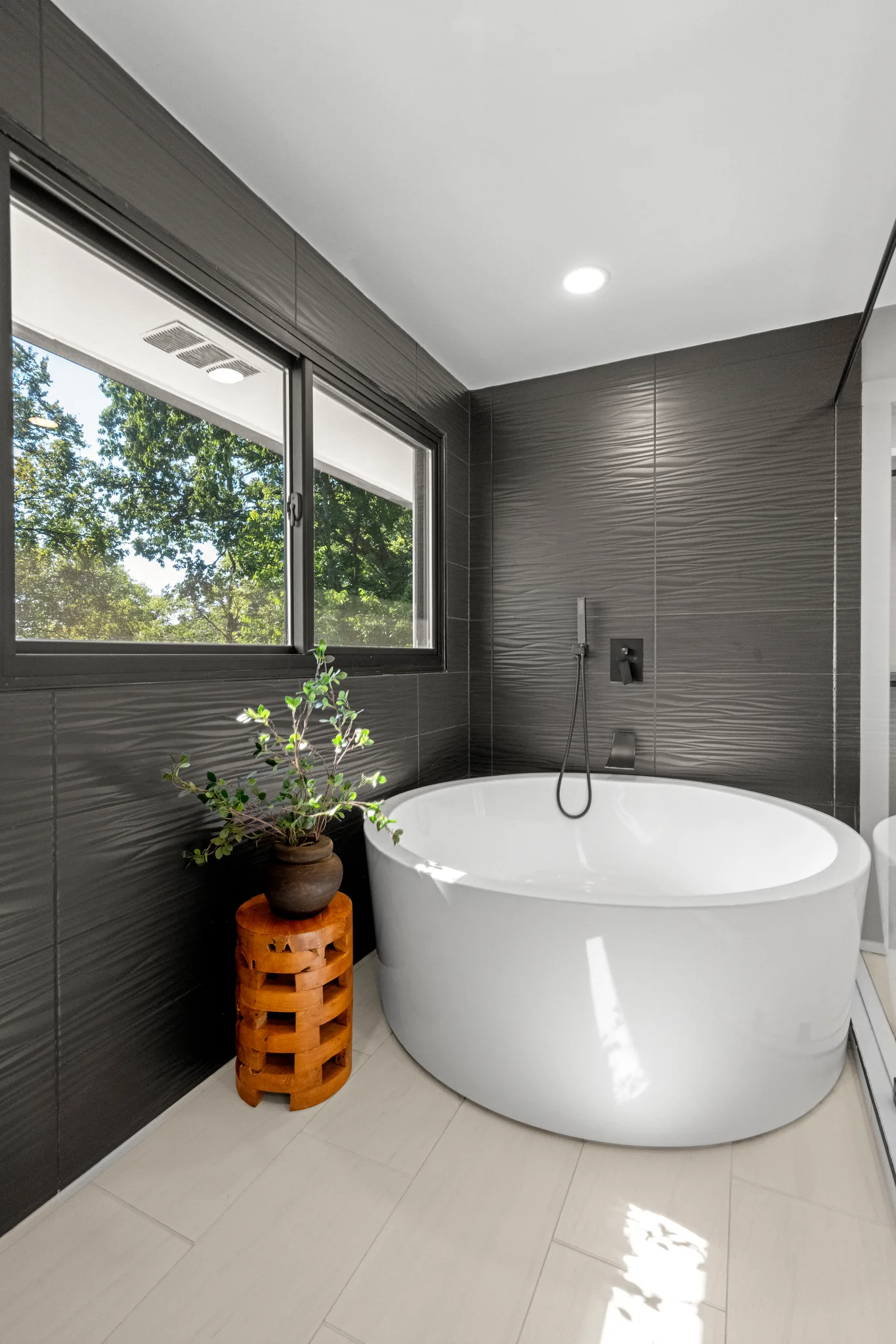 Round white freestanding soaking tub against dark textured gray wall tile with black wall-mounted filler and handheld sprayer beside a window