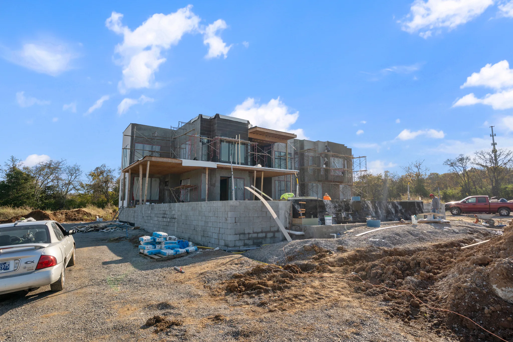 Modern two-story home under construction with scaffolding, a concrete block retaining wall, stacked material pallets and graded dirt in the foreground