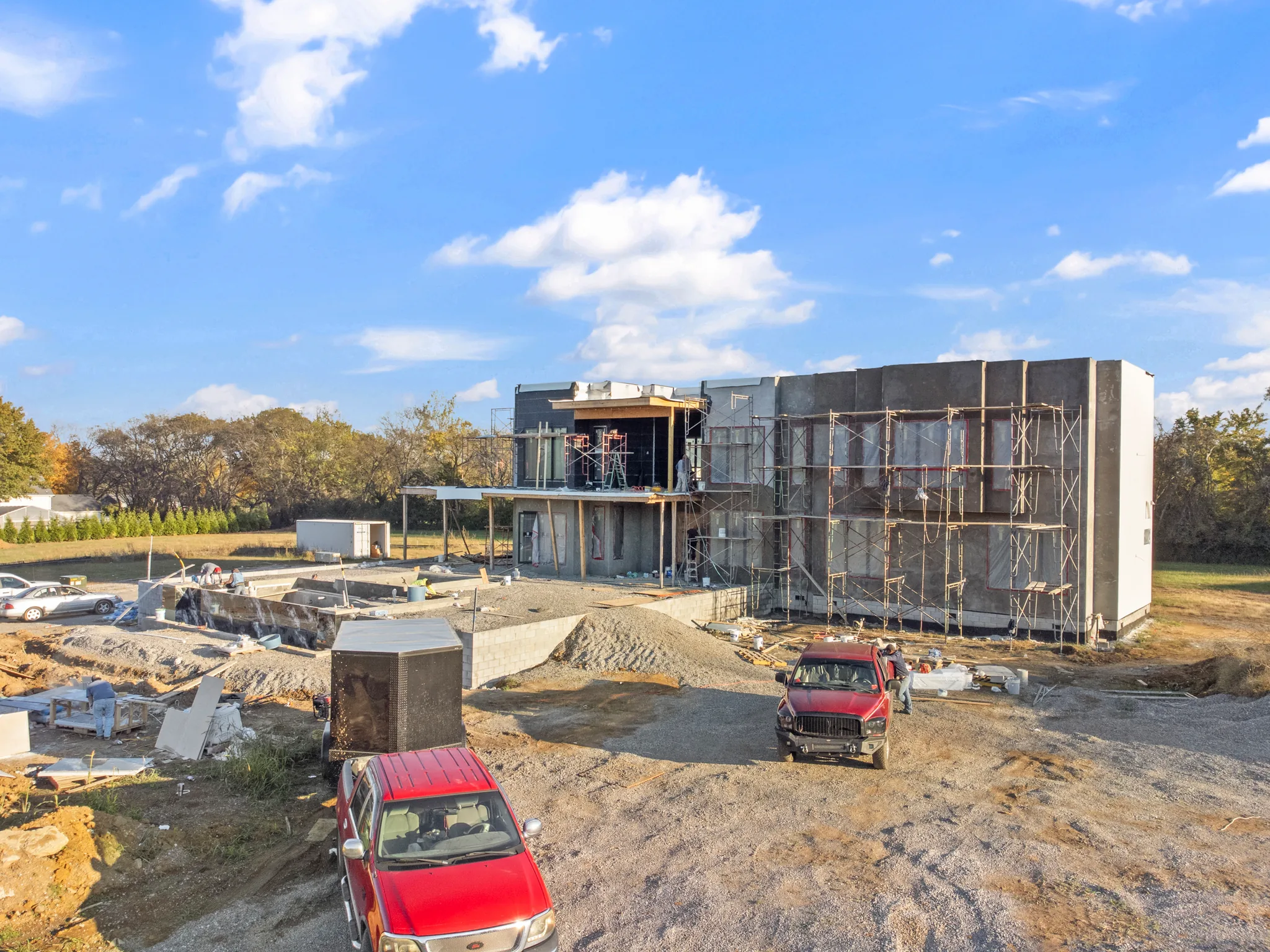 Flat-roof modern home under construction with gray panel walls, scaffolding along one wing, pickup trucks and gravel site work in the foreground