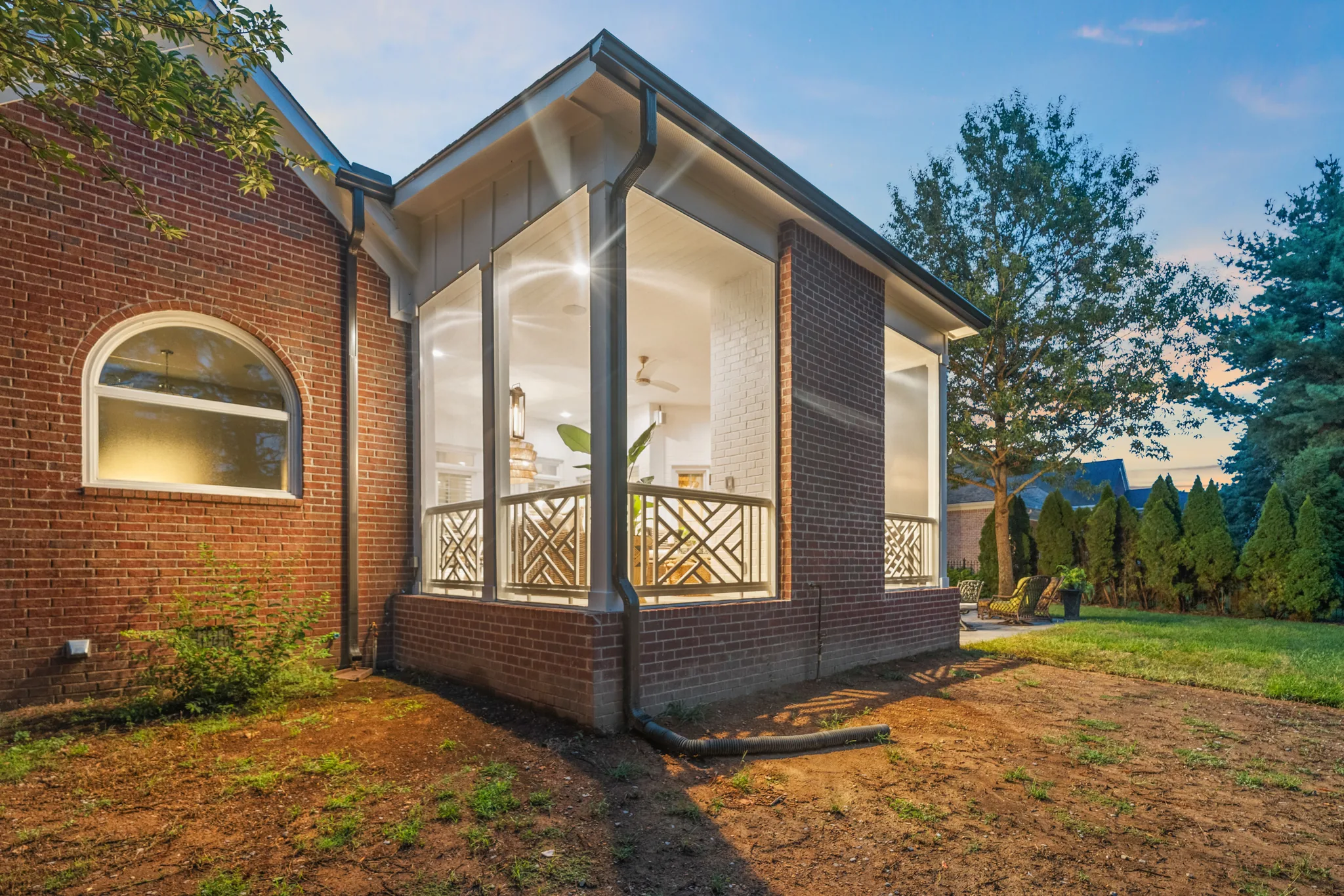 Screened porch addition with brick base, board and batten gable, Chippendale-style railing and lit interior attached to a red brick home at dusk