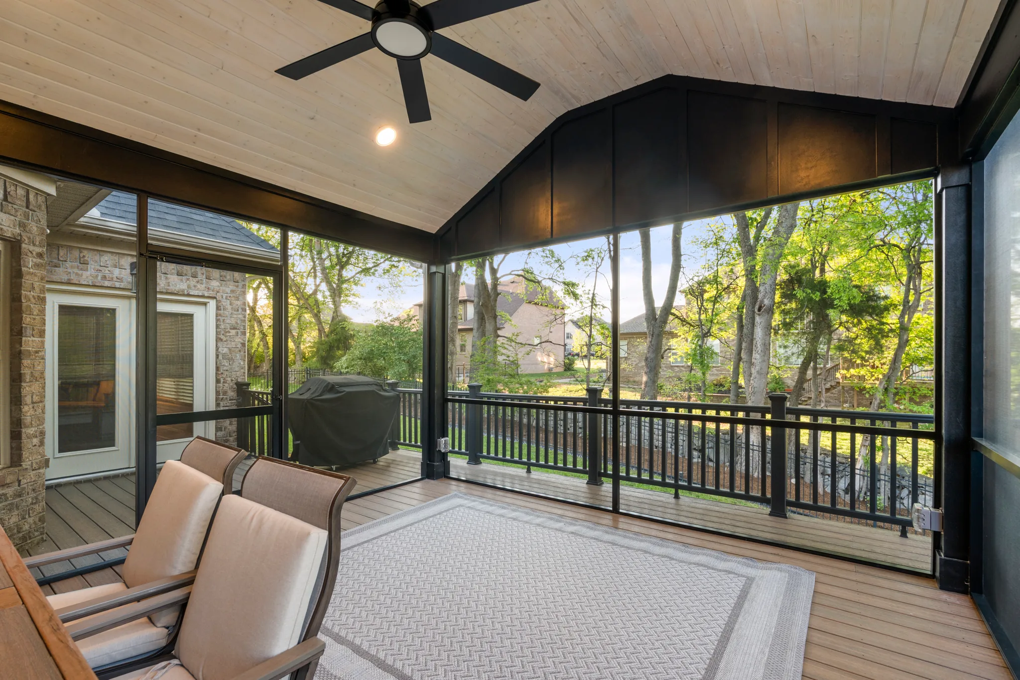 Screened porch with vaulted pine ceiling, black ceiling fan, dark board and batten gable, composite floor, patio chairs and an area rug