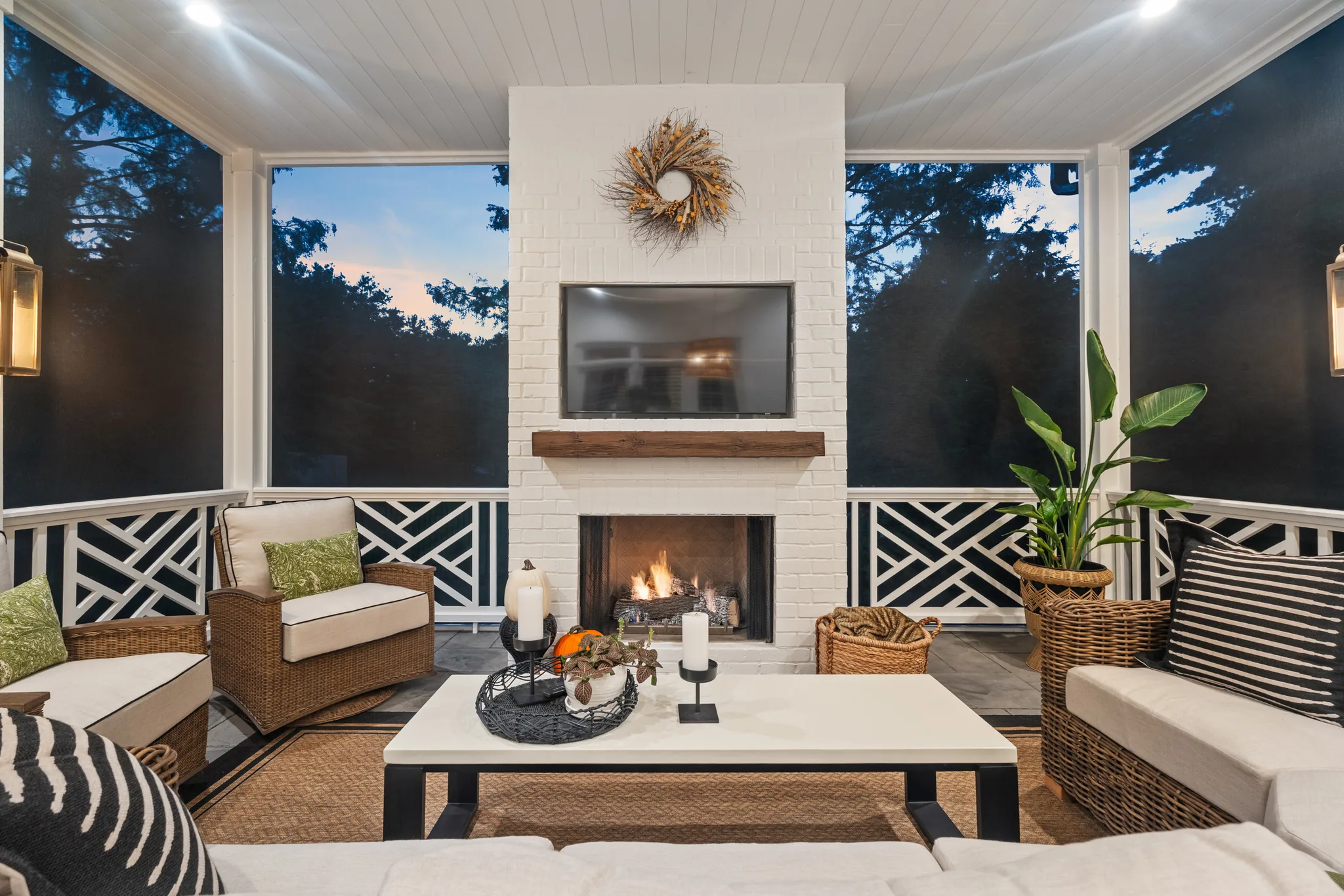 Screened porch at dusk with a white painted brick fireplace, wood mantel, mounted TV, black Chippendale-pattern railing and wicker seating