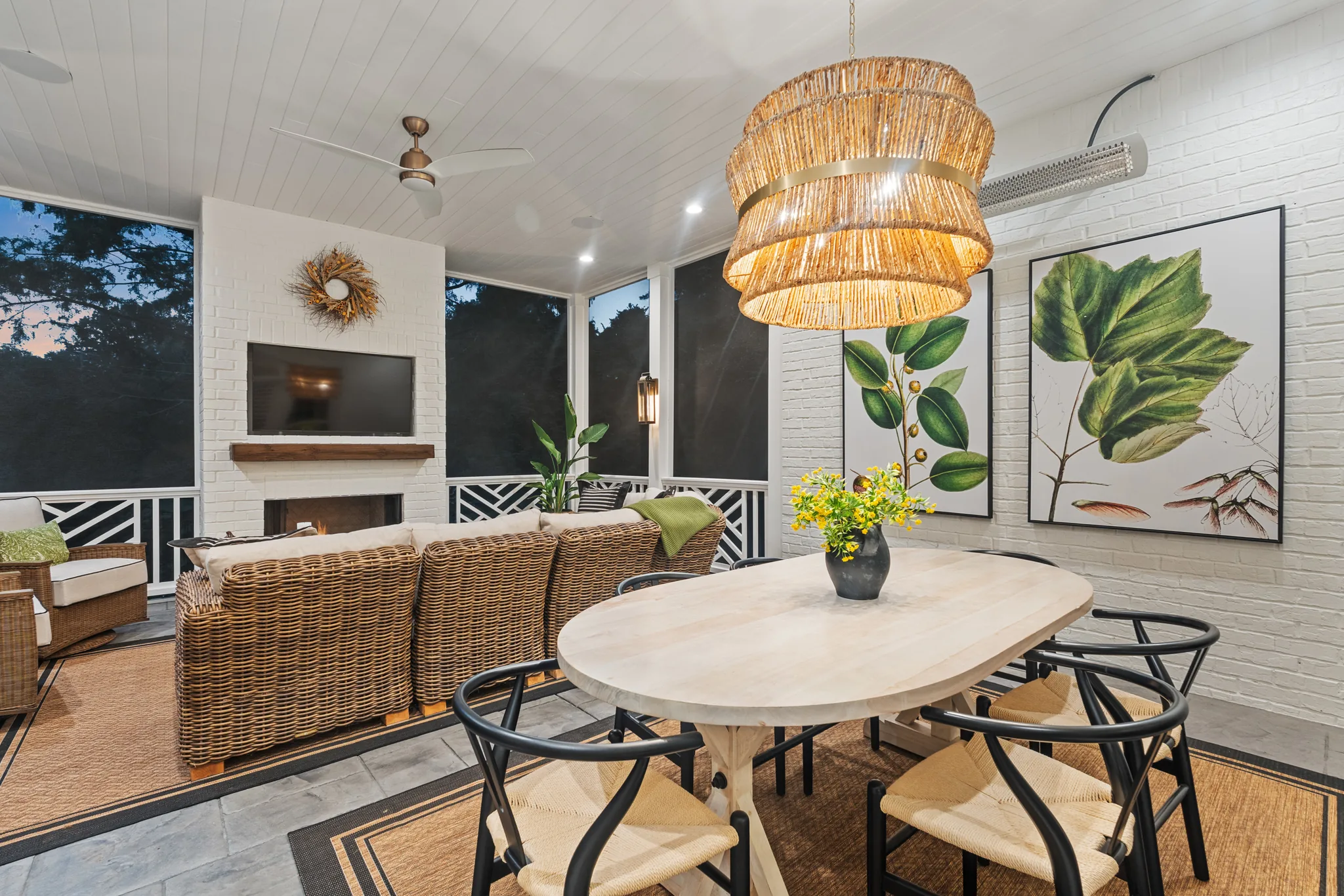 Screened porch dining area with an oval whitewashed table, black wishbone chairs, rattan drum chandelier and painted brick walls beside a wicker sectional and fireplace