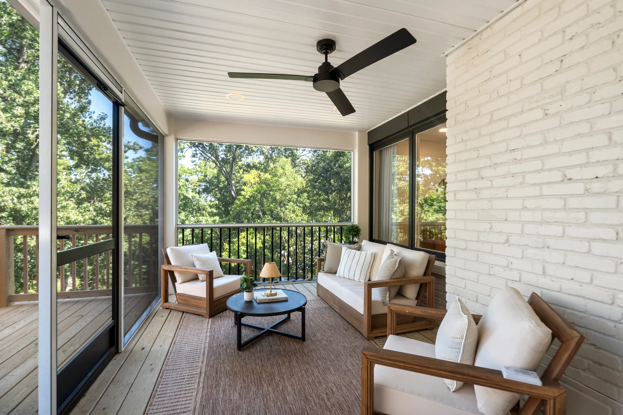 Screened porch with white beadboard ceiling, dark ceiling fan, painted white brick wall, wood furniture with cream cushions and treetop views