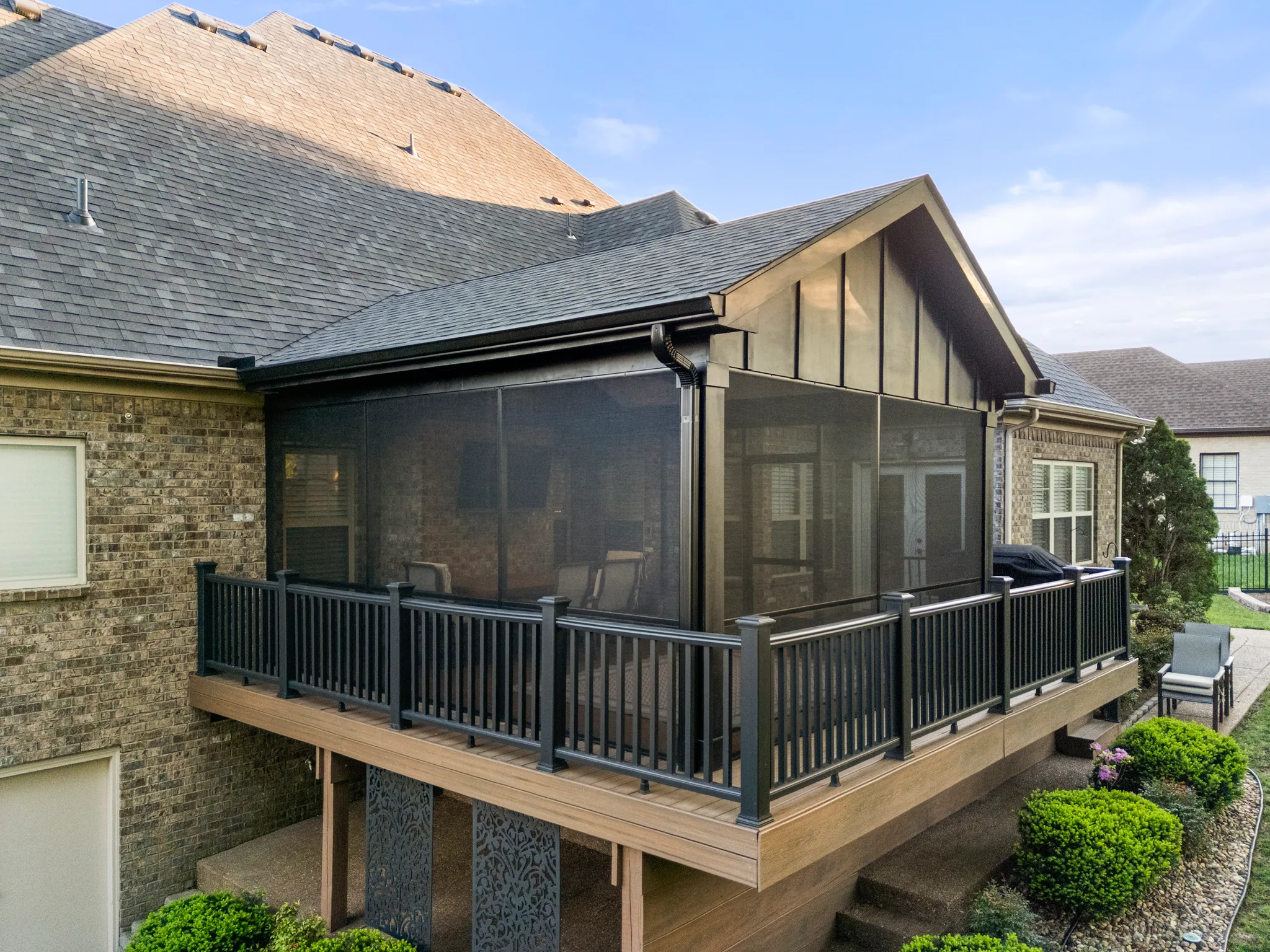 Screened porch with gable roof and shingles attached to a brick home, joined to a raised composite deck with black metal railings