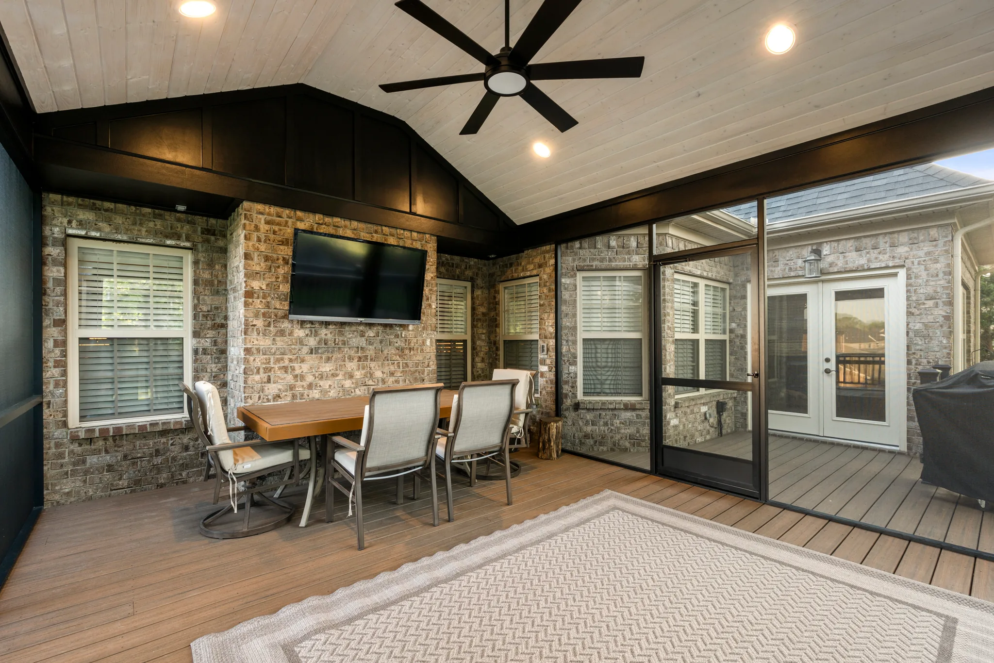 Screened porch with brick walls, whitewashed plank ceiling, dark board and batten gable, wall-mounted TV, patio dining set and composite decking