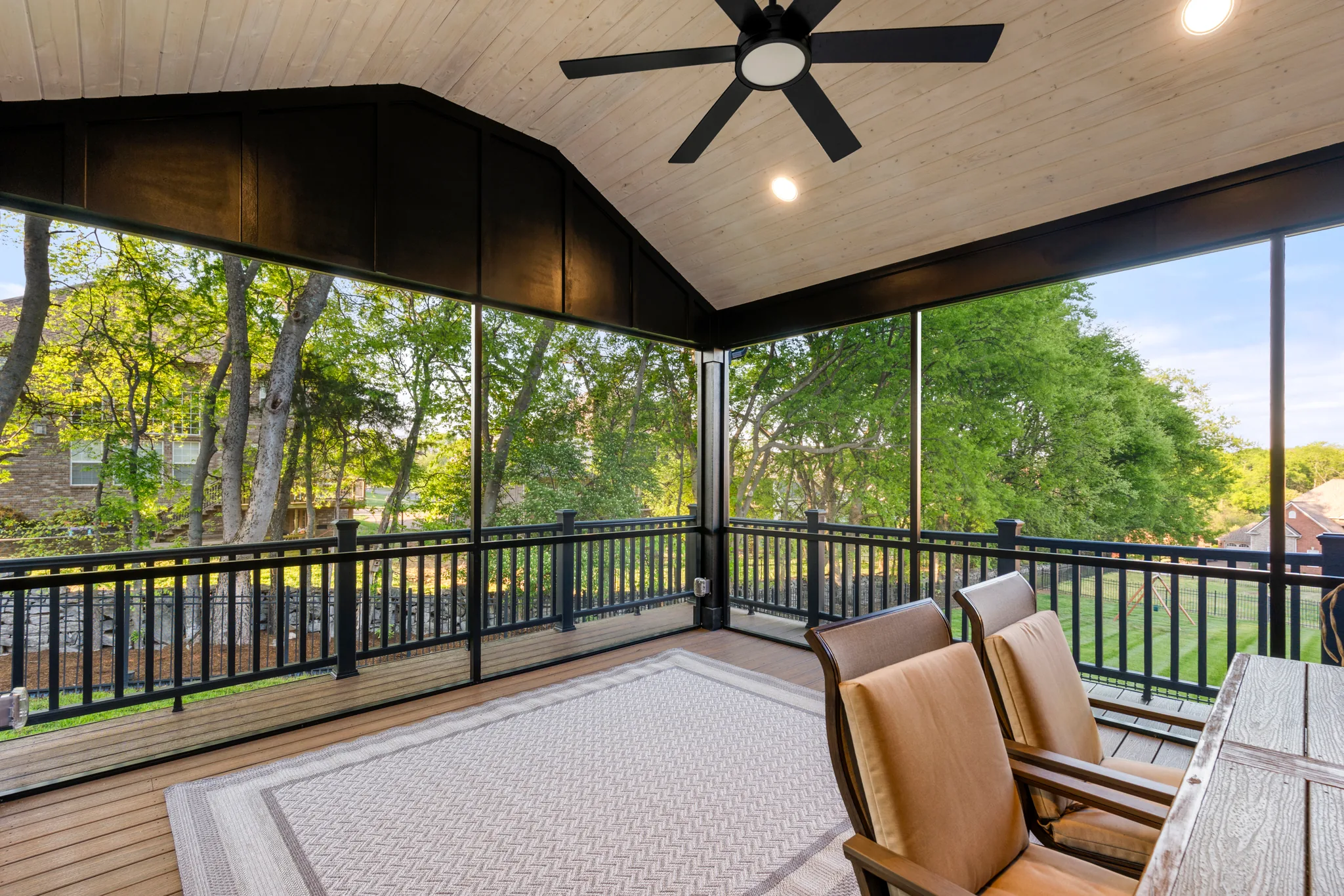 Screened porch with a vaulted whitewashed wood ceiling, black ceiling fan, black metal railing and an outdoor rug over composite decking