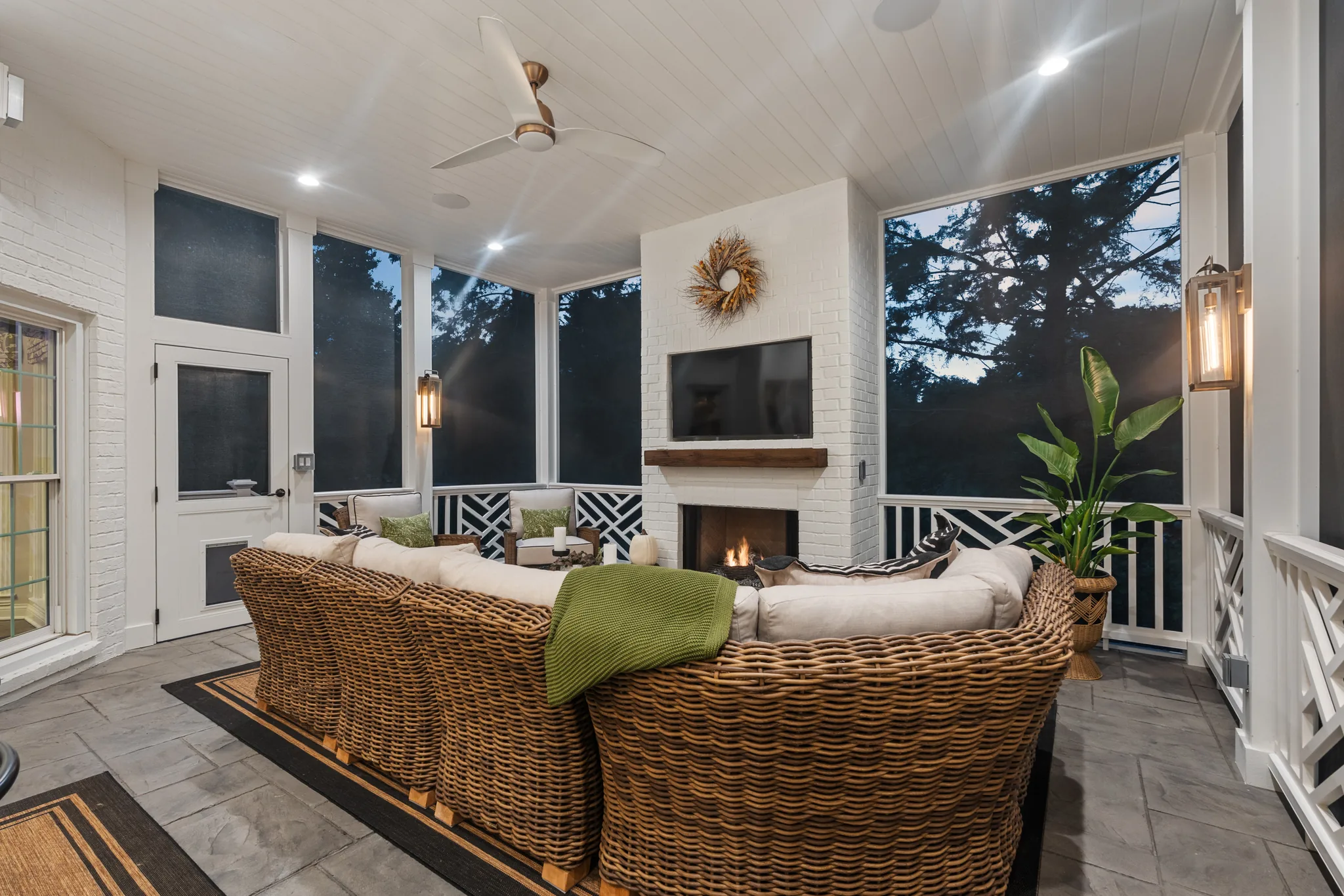 Screened porch with painted brick fireplace, mounted TV, wicker sectional, tile floor, white plank ceiling and dark lattice railing panels
