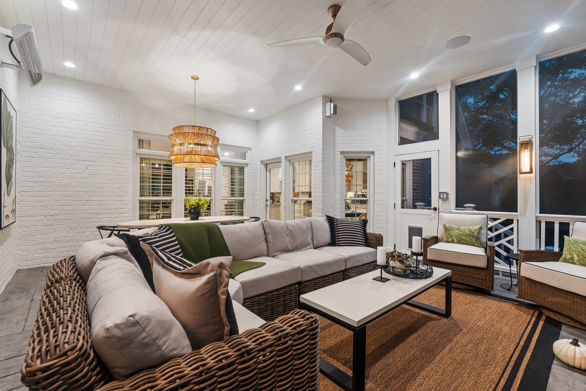 Screened porch with painted white brick walls, white plank ceiling, wicker sectional with gray cushions, woven chandelier and ceiling fan at dusk