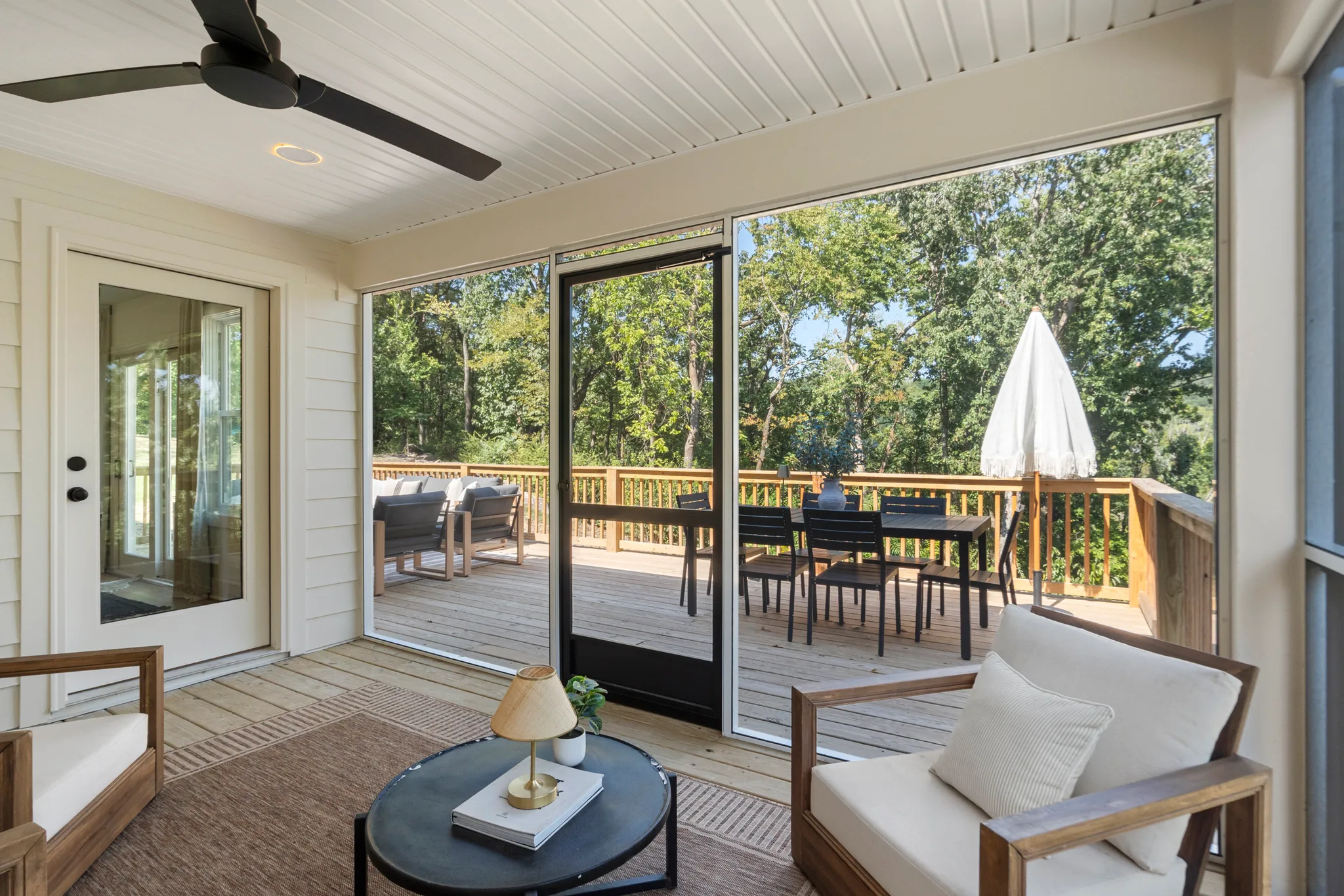 Screened porch with white beadboard ceiling, dark ceiling fan and wood furniture, looking through a screen door to a wood deck and trees