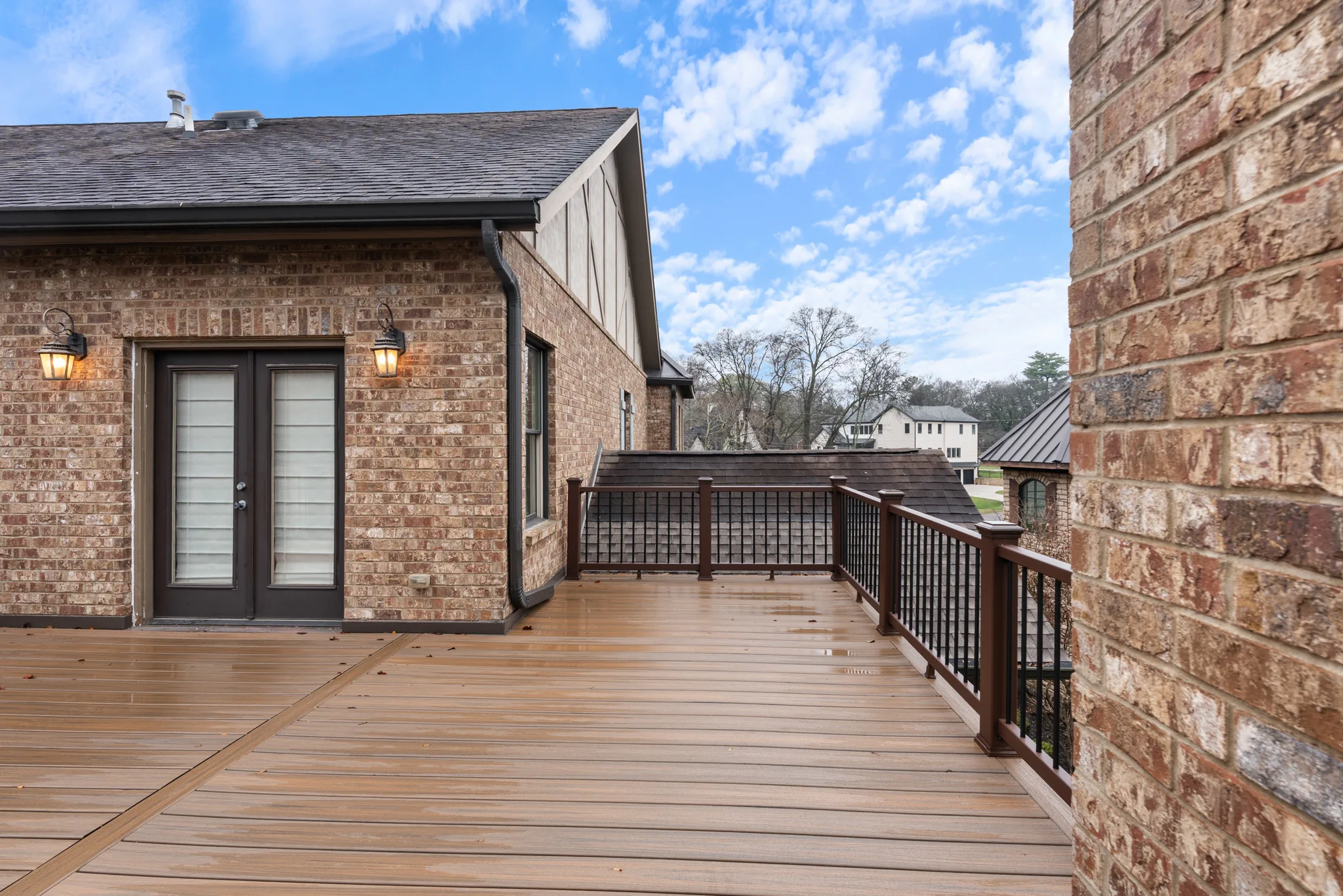 Second-story composite deck on a brick home with white French doors, wall lanterns, and dark railing with black metal balusters