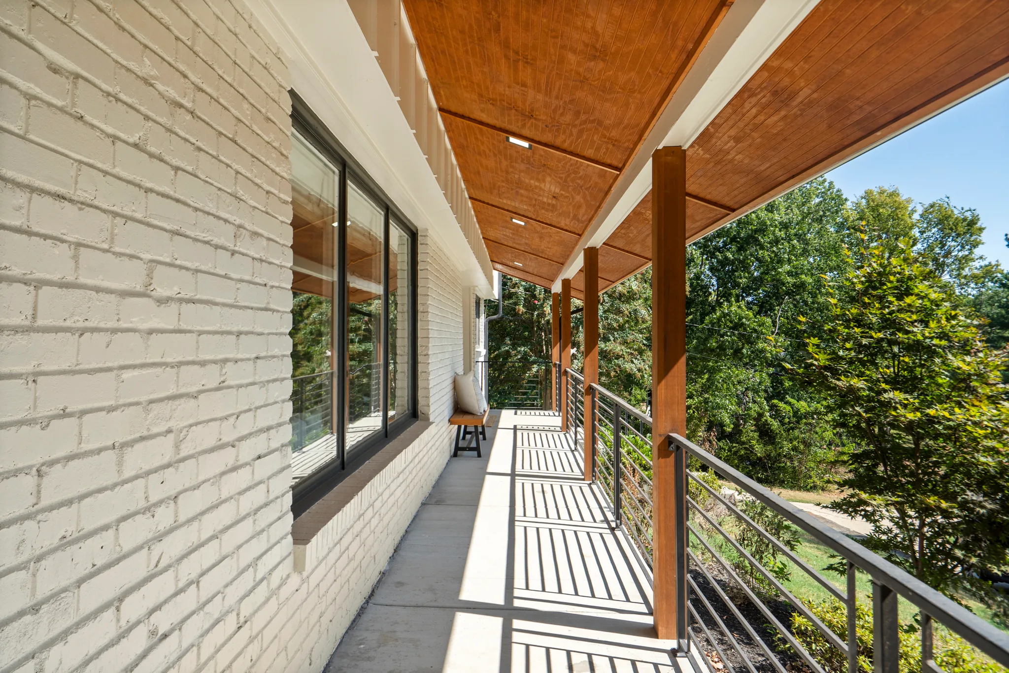 Narrow covered side porch with white painted brick wall, stained wood tongue-and-groove ceiling, wood posts, black metal cable railing, and trees beyond