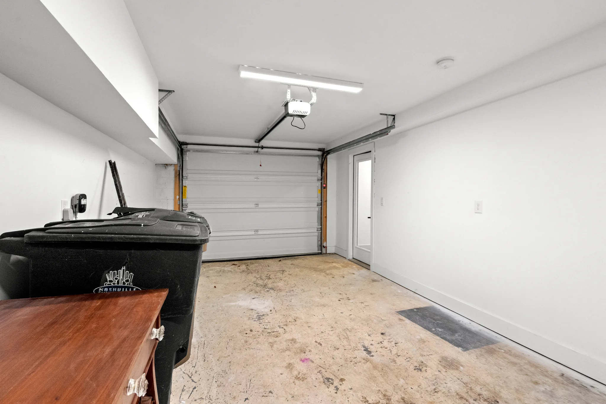 Single-car garage interior with white painted walls, sectional overhead door, concrete floor, linear ceiling light and a glazed side entry door