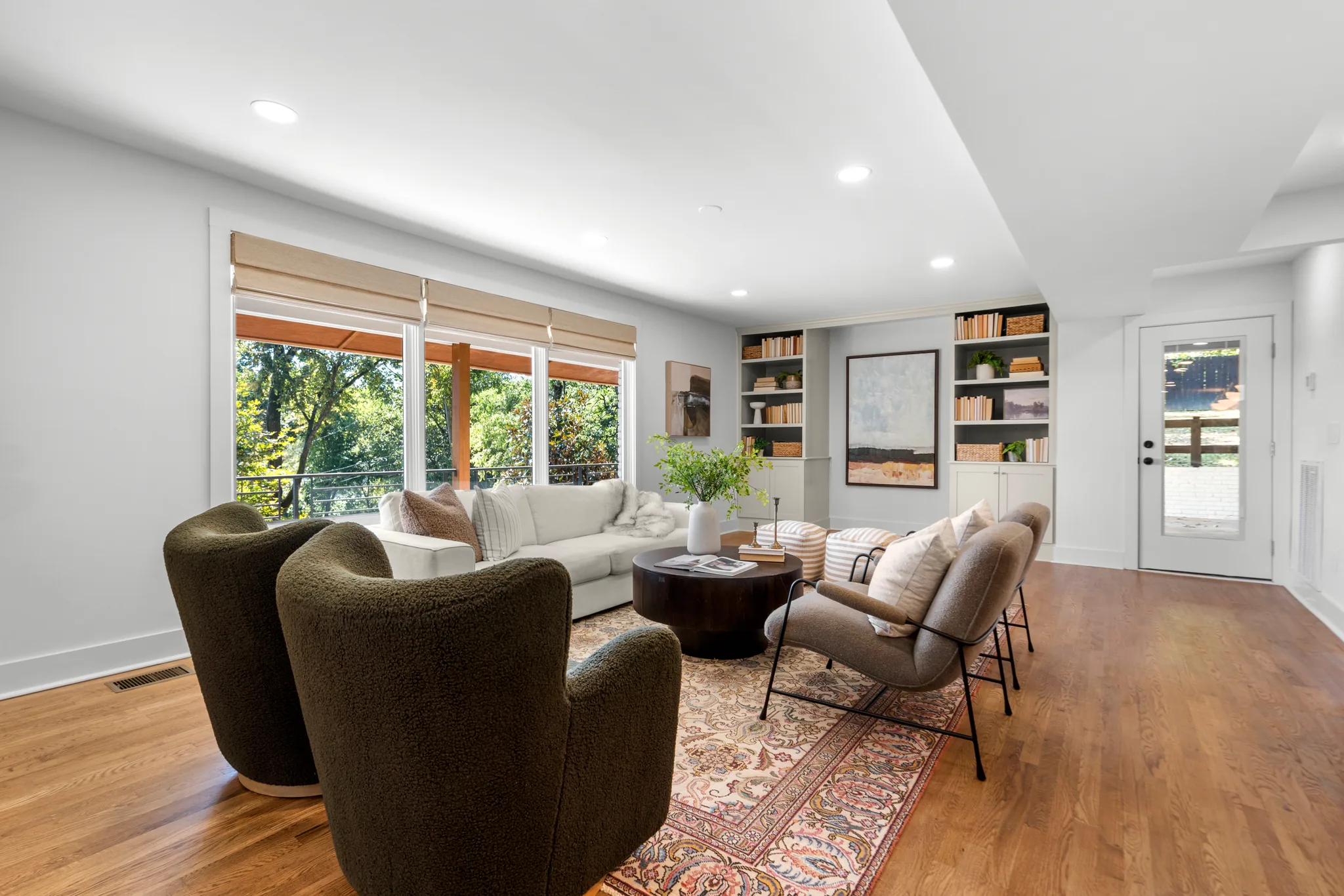 Renovated sitting area with light gray walls, oak floors, dark built-in shelves, woven shades and a glass door to a deck