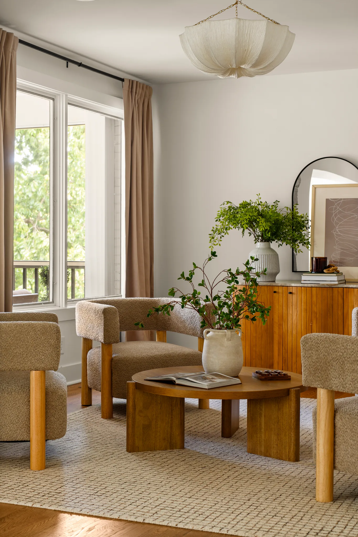 Sitting room with boucle armchairs around a round wood coffee table, woven rug, tall window with linen curtains, and a wood sideboard