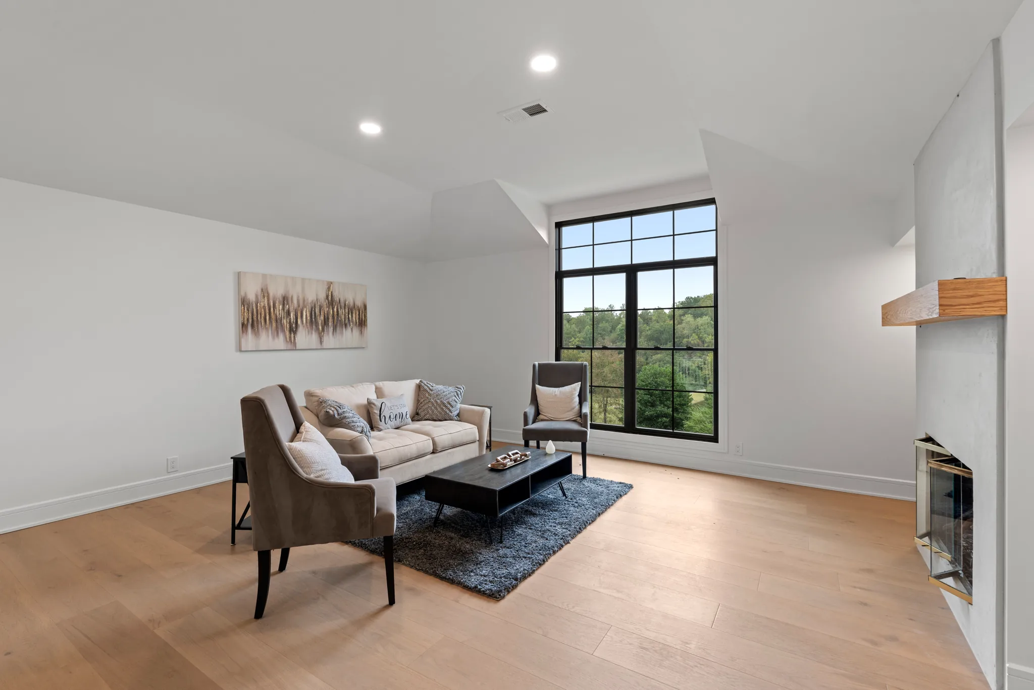 Sitting room with white walls, wide oak flooring, a tall black grid window and a plaster fireplace with a wood mantel shelf
