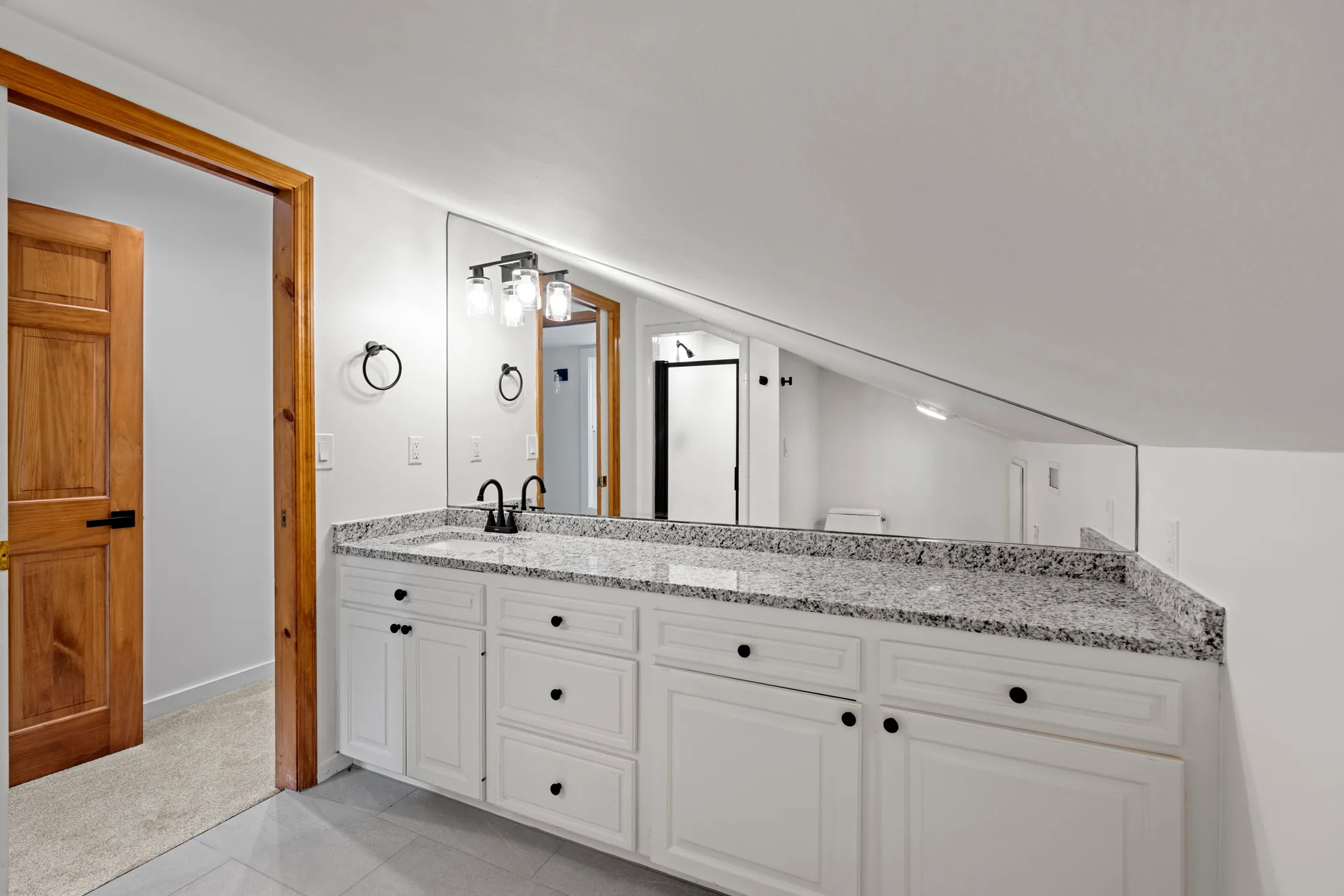 Bathroom vanity with white raised-panel cabinets and speckled granite counter set under a sloped ceiling with a large frameless mirror