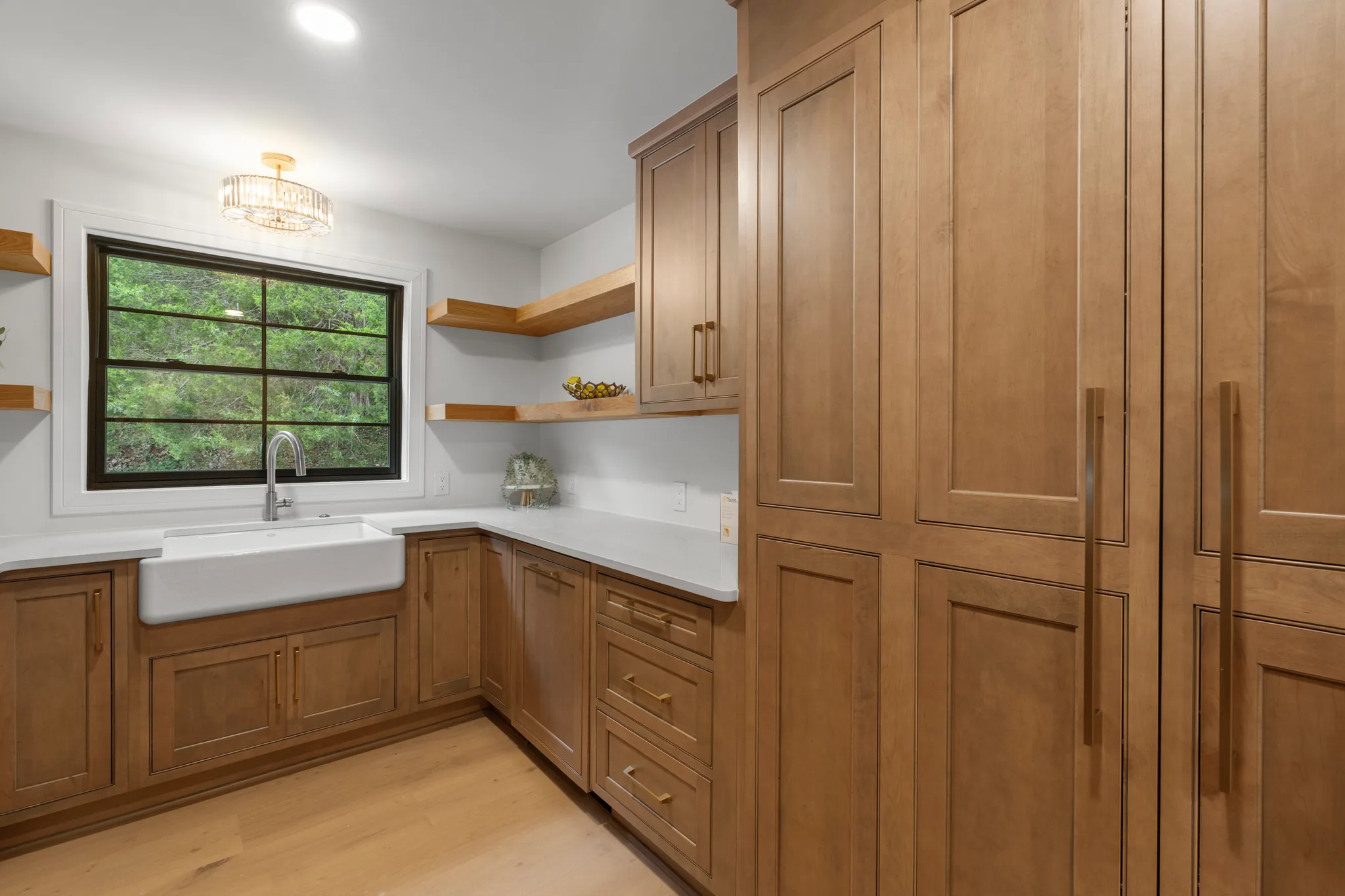 Kitchen area with medium stained wood cabinets, white quartz counters, apron-front farmhouse sink under a black-framed window, and floating wood shelves