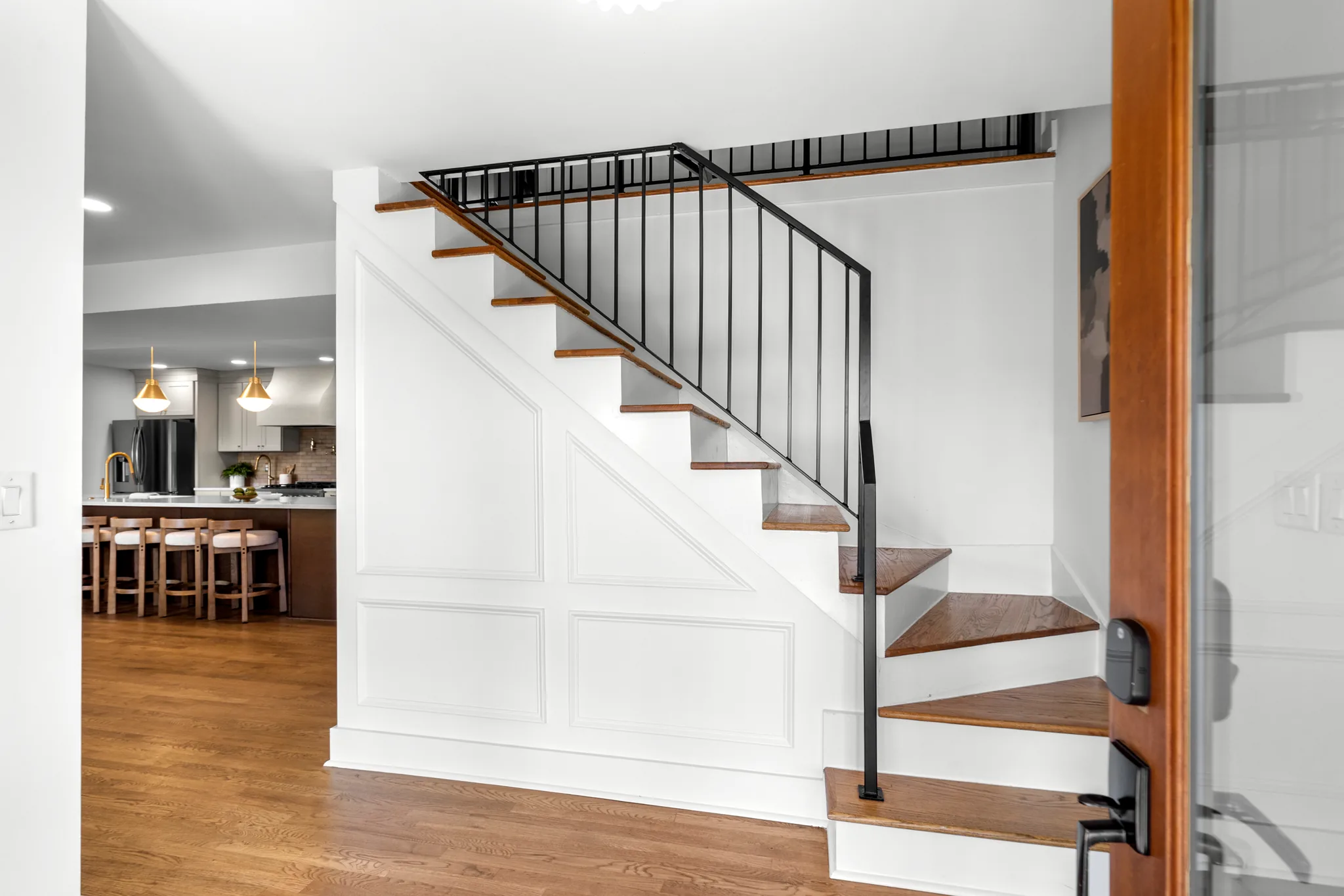Interior staircase with stained wood treads, white risers and a black metal spindle railing beside a paneled wall, with a kitchen visible beyond