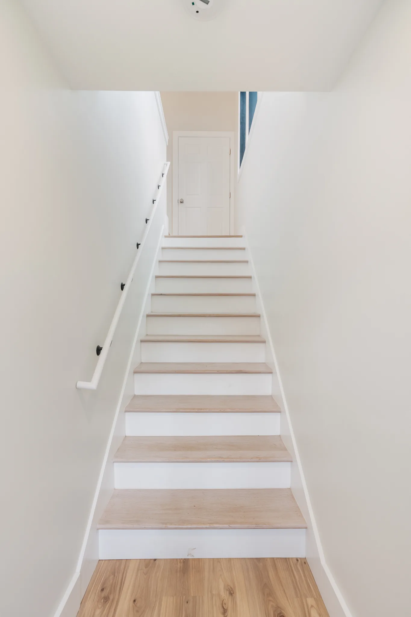 Narrow staircase with light wood treads, white risers and a white wall-mounted handrail leading up to a six-panel door