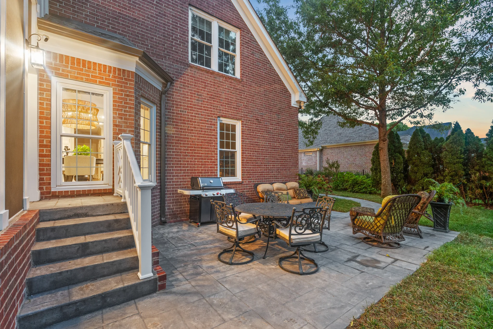 Stamped concrete patio beside a red brick home at dusk with wrought iron dining set, grill and wicker lounge chairs