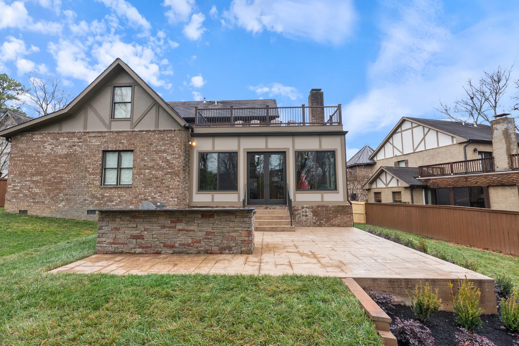 Rear of a brick and stucco home with a stamped concrete patio, stone-faced bar with grill, steps to French doors, and a railed rooftop deck