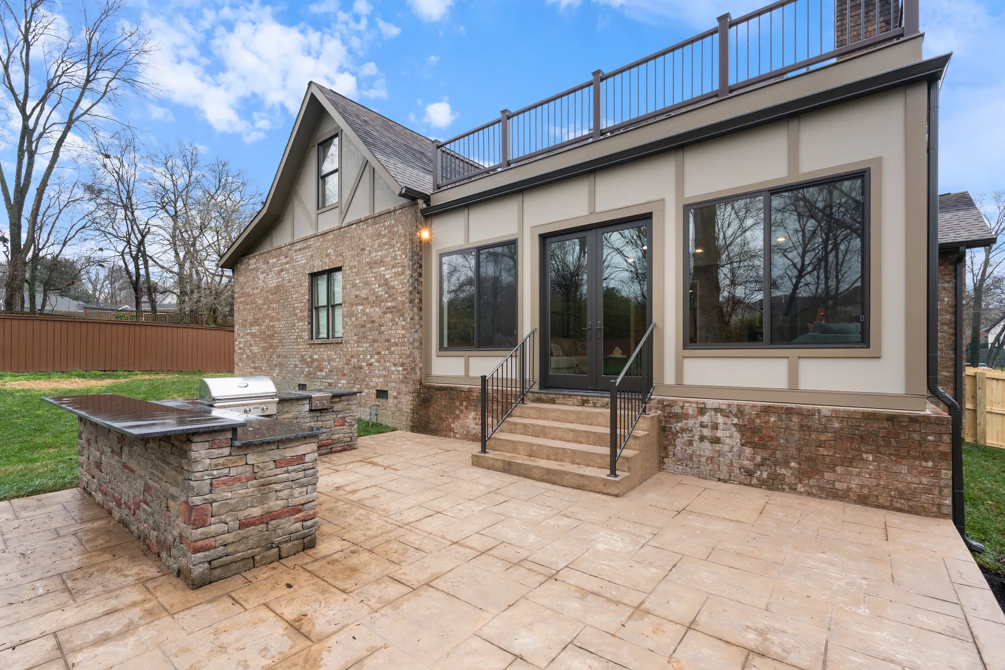 Stamped stone-look patio behind a home with a stacked stone grill island, built-in stainless grill, dark counters, and steps to French doors
