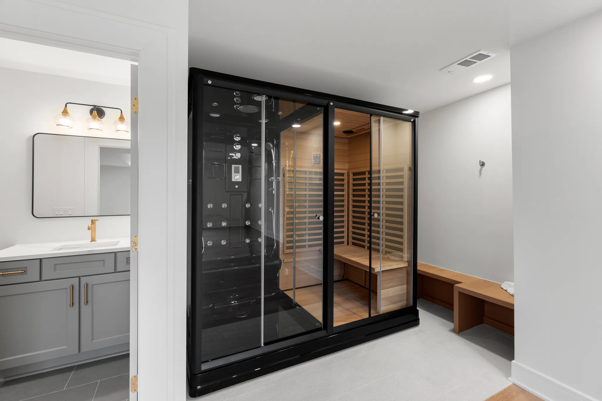 Spa room with a black steam shower unit and glass-front cedar sauna beside a wood bench and a gray vanity in the adjoining bath