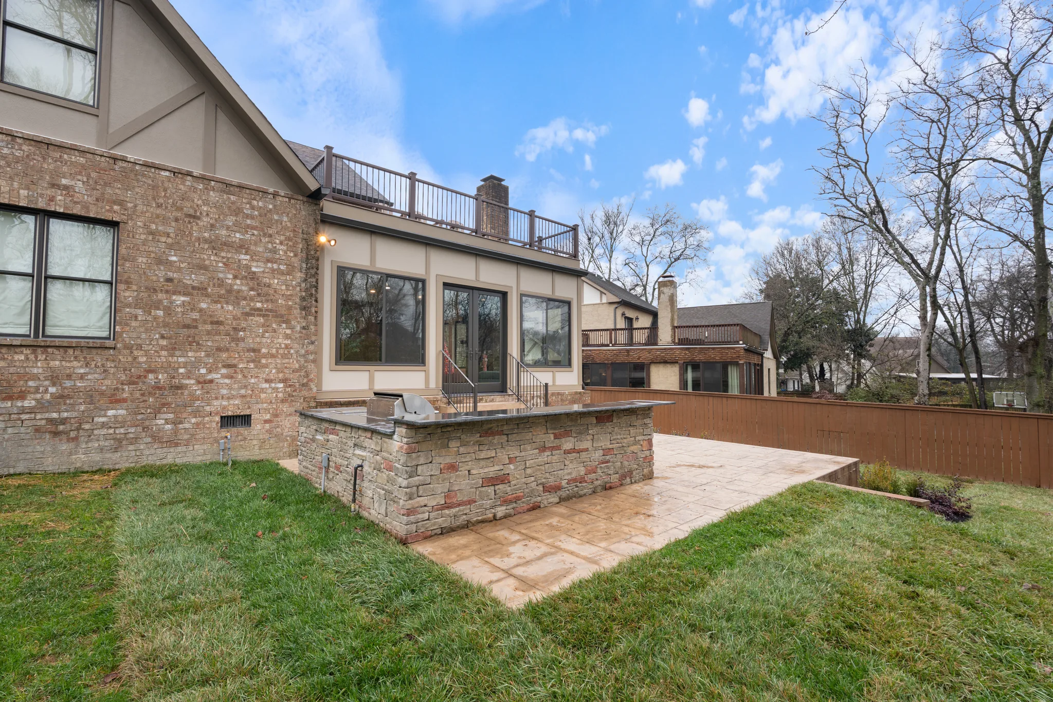 Stone-faced outdoor bar with built-in grill and stone countertop on a stamped concrete patio beside a brick home with upper deck