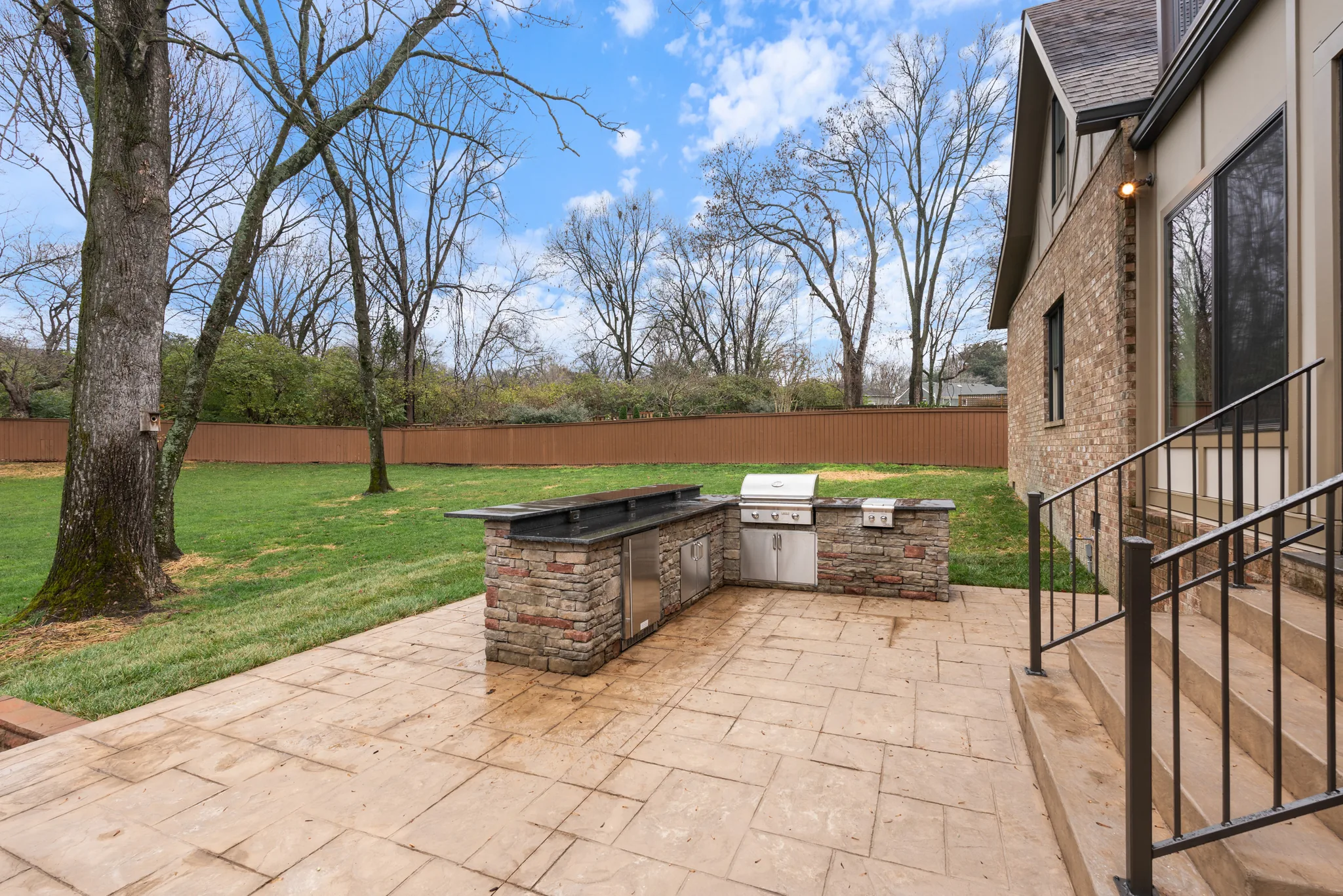 L-shaped stone outdoor kitchen with built-in grill and stainless doors on a stamped concrete patio beside a brick house and fenced lawn