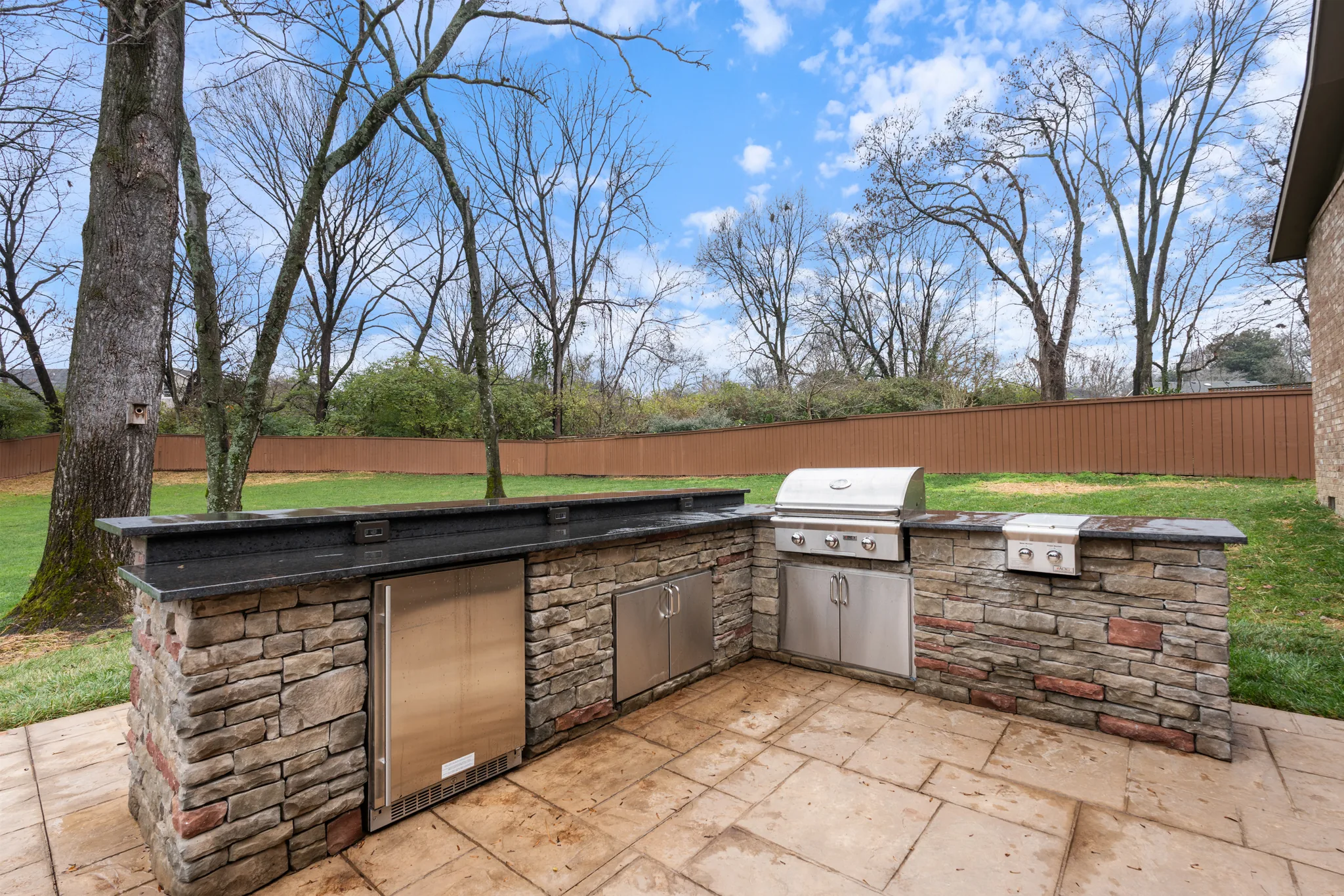 L-shaped outdoor kitchen clad in stacked stone with black granite counters, built-in grill, side burner and stainless refrigerator on a stamped concrete patio