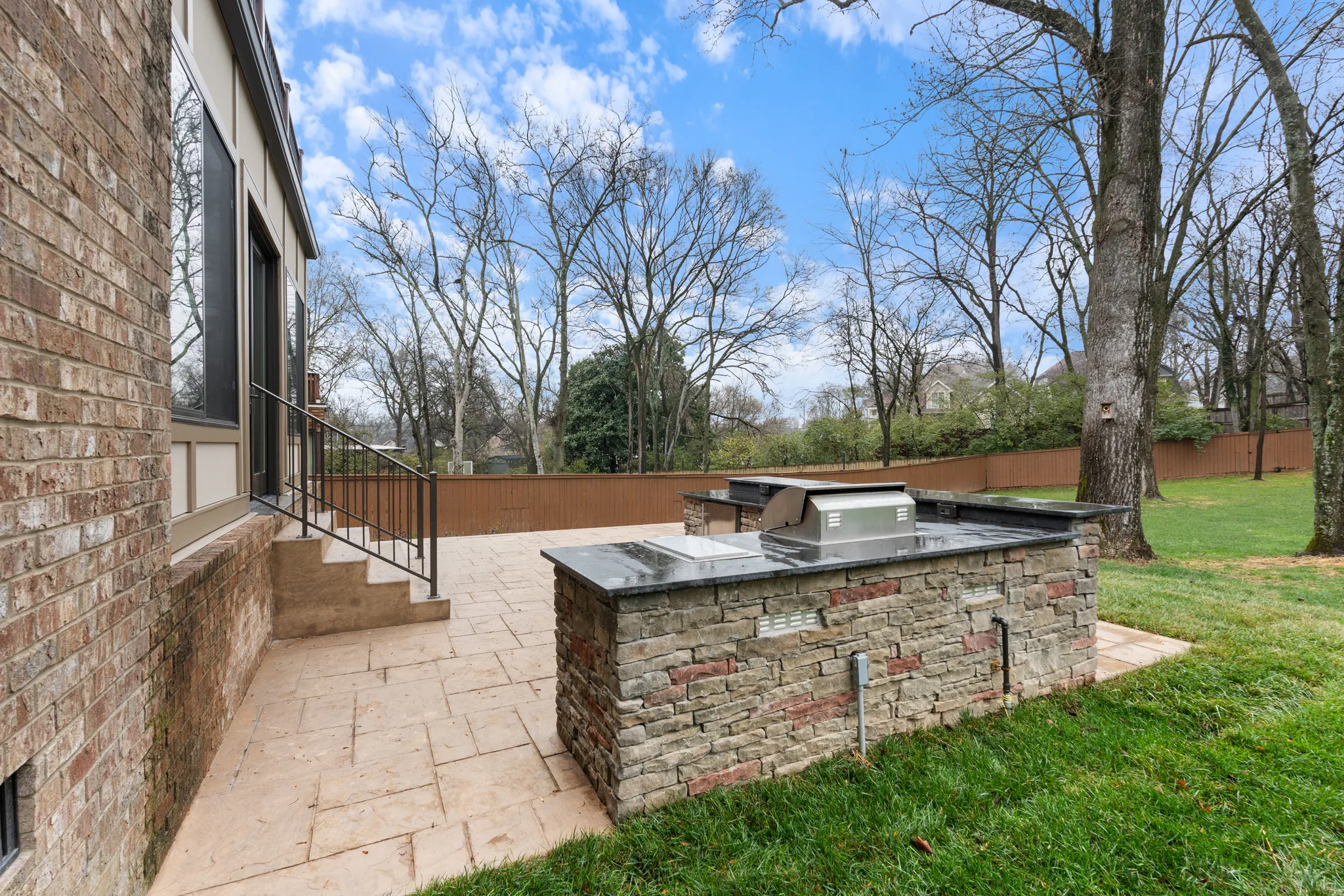 Outdoor kitchen island faced in stacked stone with dark granite counter and built-in stainless grill on a stamped concrete patio beside a brick home