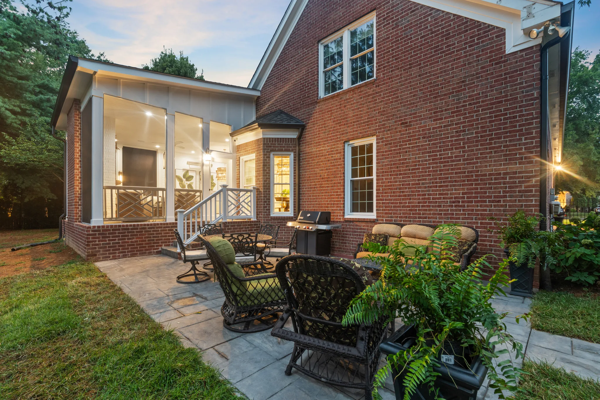 Stone paver patio with black wicker chairs and a grill beside a red brick home with a lit screened porch at dusk