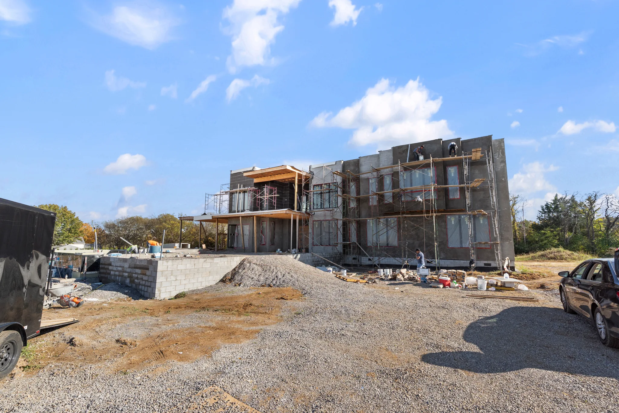 Two-story home under construction with gray stucco walls, scaffolding along the rear, covered upper deck and a gravel drive in front