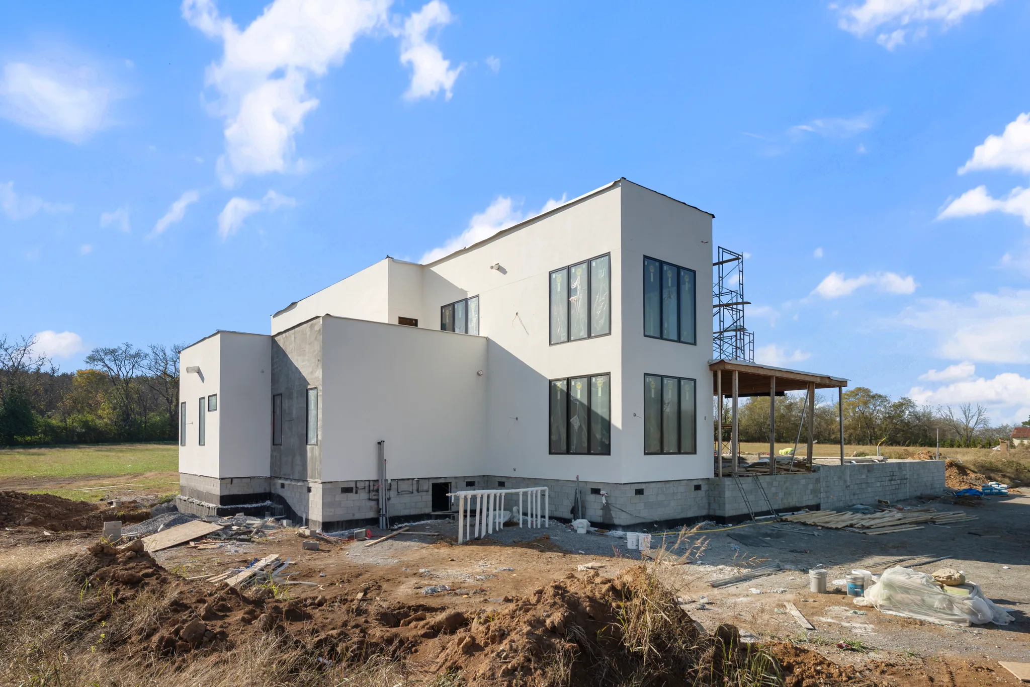 Side elevation of a white stucco modern home under construction with black-framed windows, block foundation, scaffolding and graded dirt in the foreground